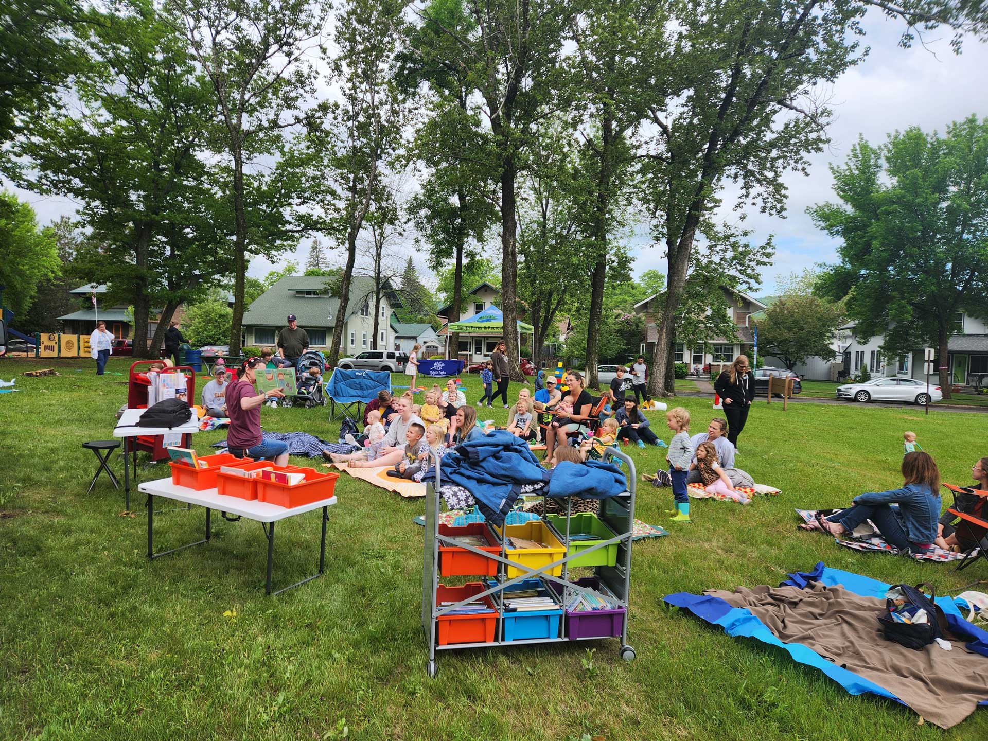 A group of people, including families and children, are enjoying a picnic in a park near the Duluth Public Library. Several blankets are spread out on the grass, and there are tables with bins and a colorful storage cart nearby. Trees and houses are visible in the background.