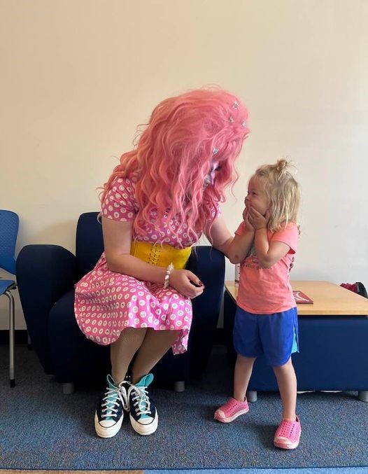 A person with long pink hair wearing a colorful dress and black sneakers sits on a chair at the Duluth Public Library, leaning forward to talk with a young child. The child stands and smiles, touching their own face with both hands. They both appear to be in a cheerful mood.