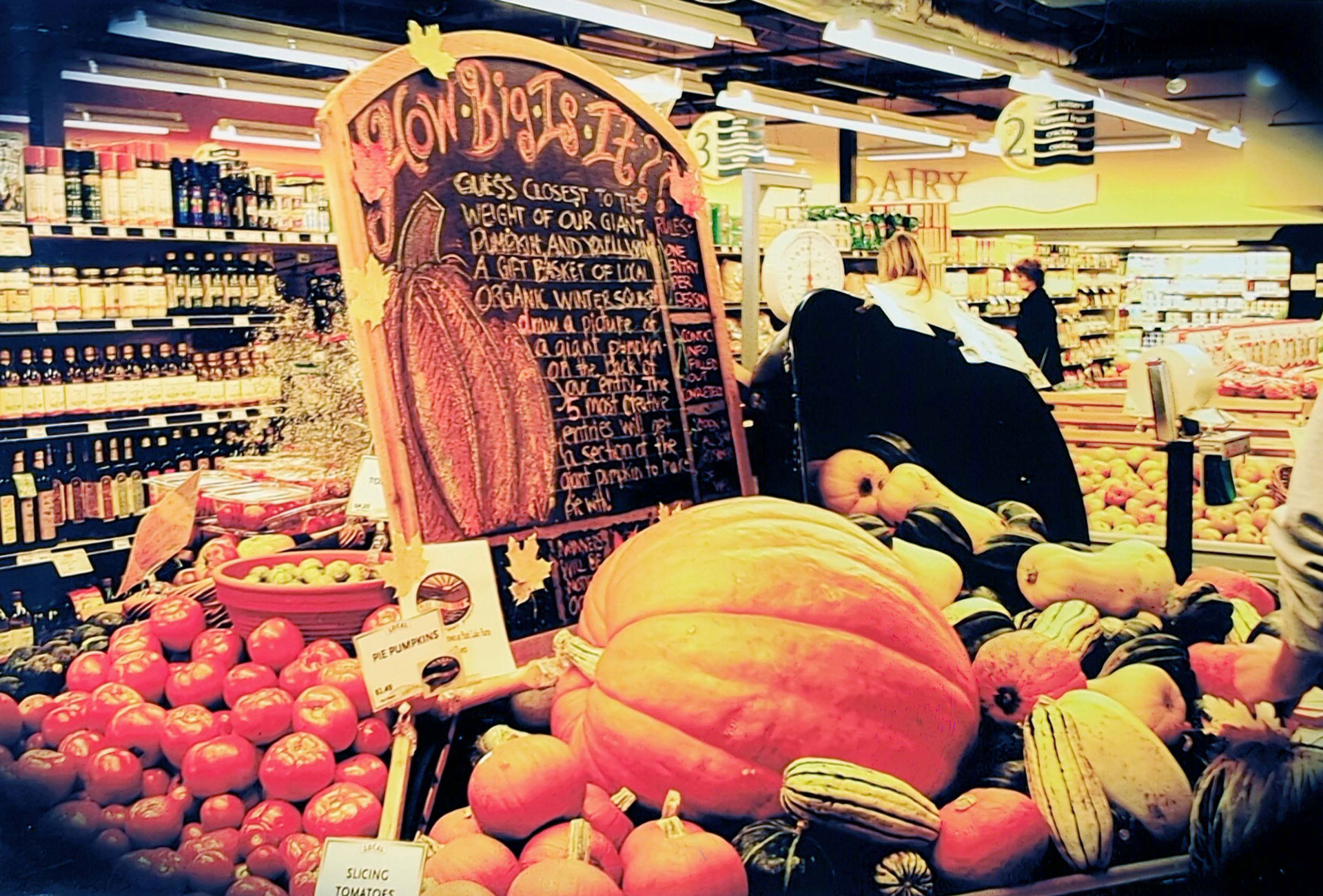 A charming display at the Whole Foods Coop Duluth MN features various pumpkins, squash, and gourds. A large pumpkin is surrounded by smaller produce. A blackboard sign invites customers to guess the weight of the giant pumpkin. Shelves of canned and bottled goods are in the background.