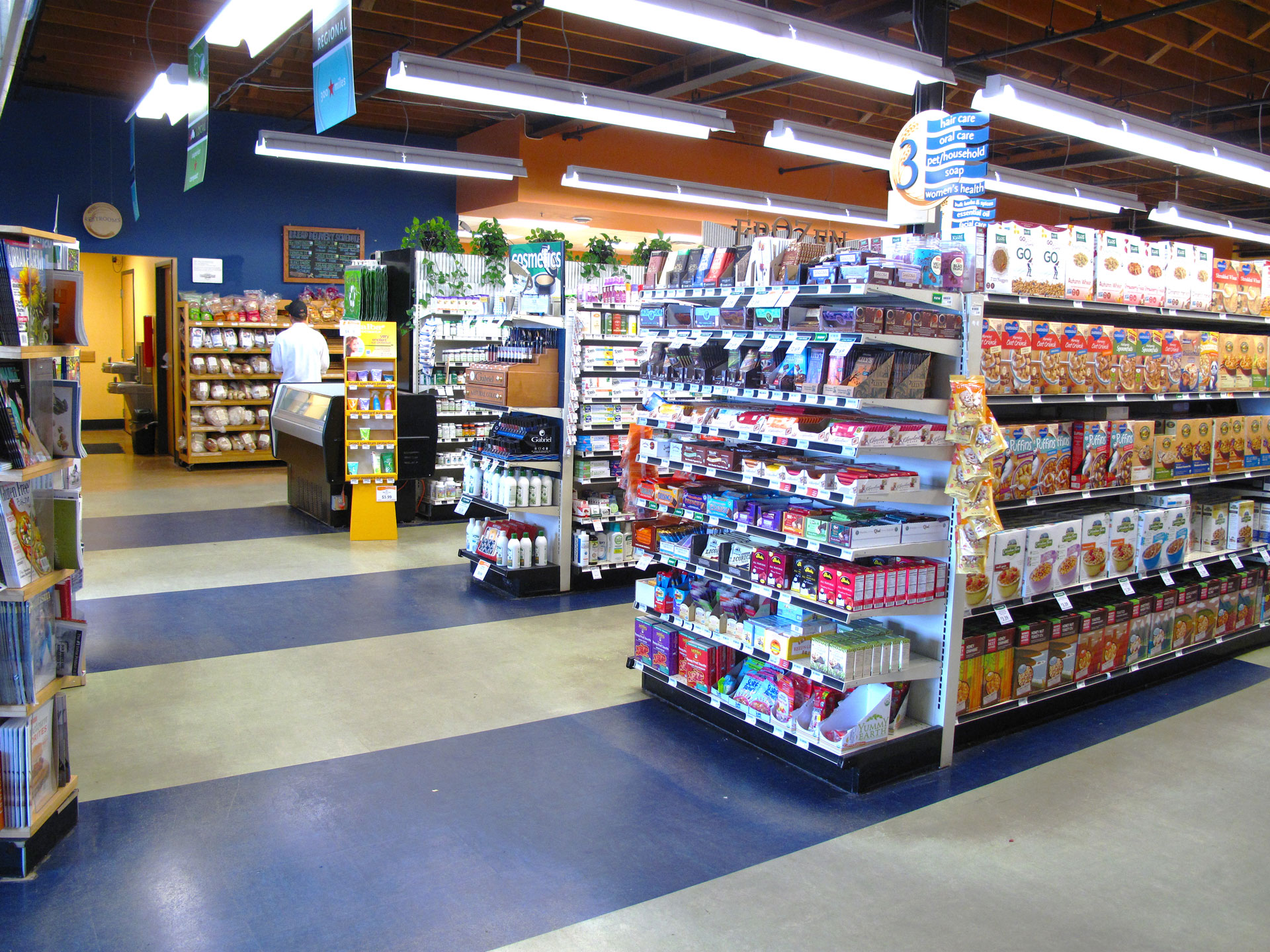 A grocery store aisle at Whole Foods Coop Duluth MN with shelves stocked with various food items, cereals, and household products. The aisle is clean and well-lit with signs for different sections. A person in the distance appears to be working or shopping near the checkout area.
