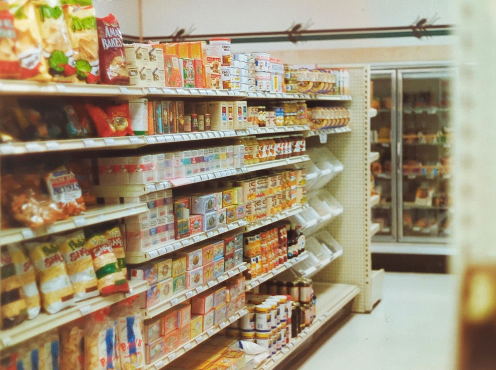 A grocery store aisle with shelves stocked with various packaged foods and snacks. The left side has chips and snack bags, while the middle shelves display cereal boxes and assorted food items. Refrigerated goods are visible in coolers on the right side, reminiscent of Whole Foods Coop Duluth MN.