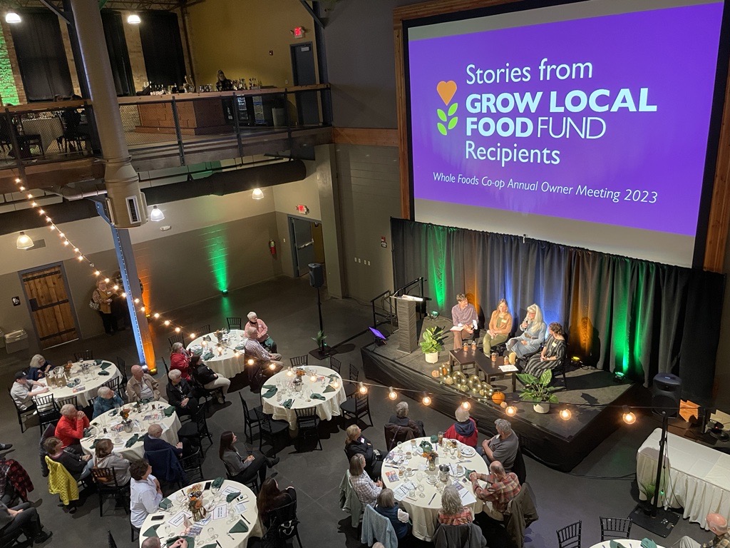 A large room with round tables and people seated, watching a panel discussion on stage. A screen behind the panel reads, "Stories from Grow Local Food Fund Recipients" at the Whole Foods Co-op Duluth MN Annual Owner Meeting 2023. String lights hang from the ceiling.