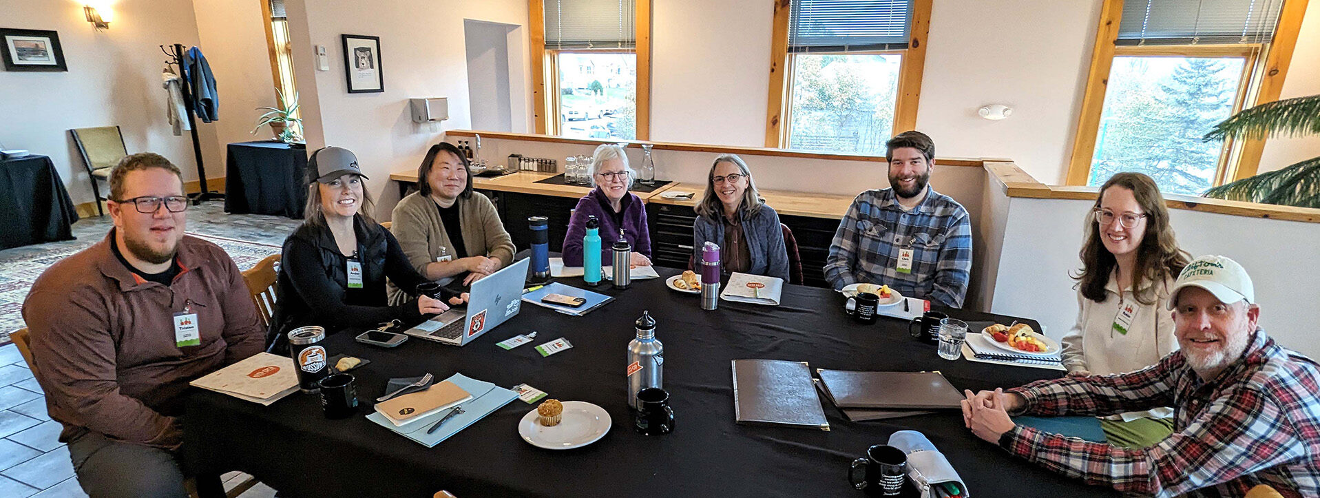A group of eight people is sitting around a rectangular table in a well-lit room. They appear to be in a meeting, with laptops, notebooks, and drinks on the table. The attendees are smiling and engaged. Large windows in the background allow natural light to illuminate the space.