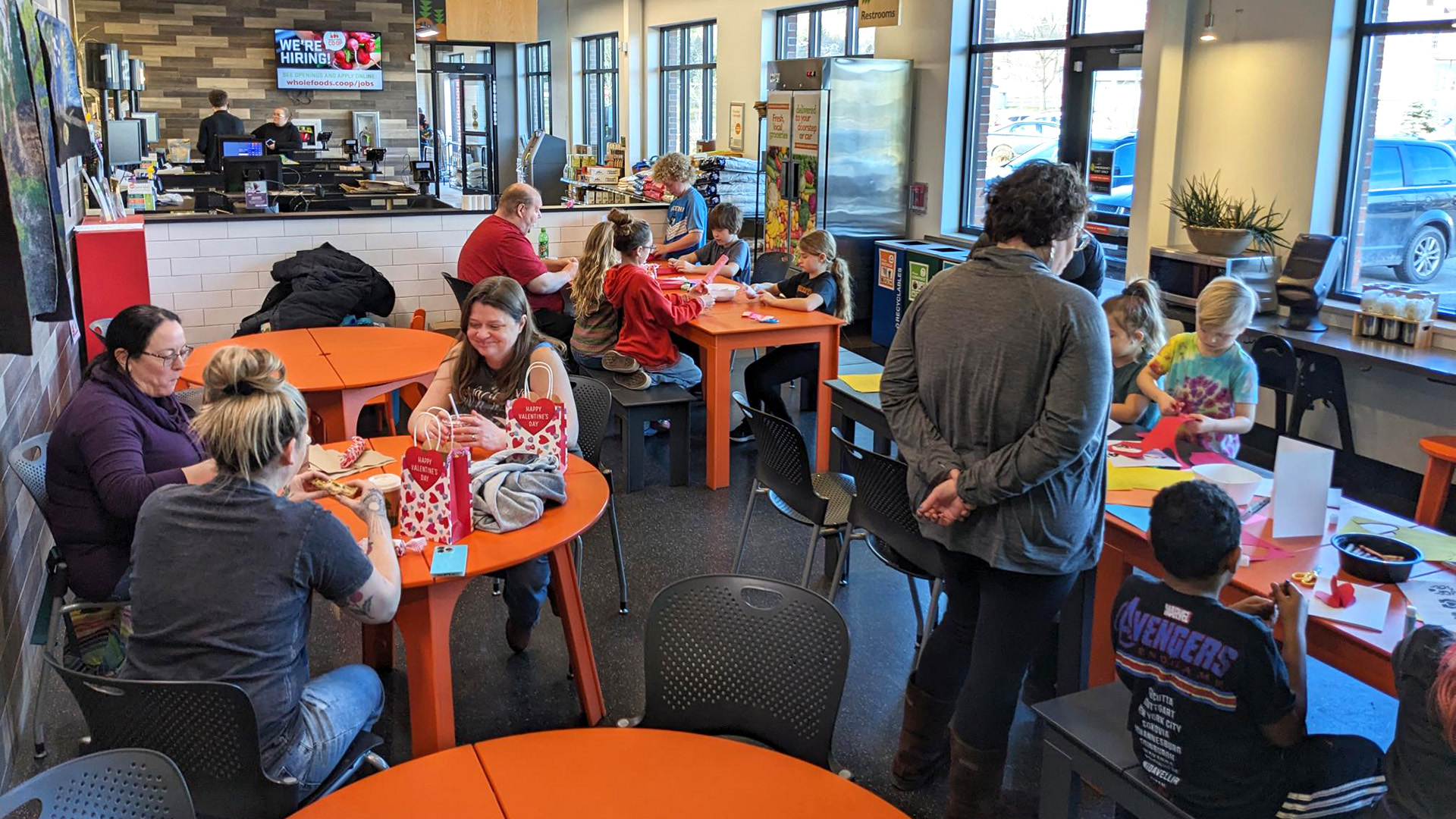 People are sitting at orange tables in a cafe at Whole Foods Coop Duluth MN, engaging in various activities such as drawing and crafting. A man is working at a counter in the background, and large windows line the walls, letting in natural light. A TV screen is visible near the counter.
