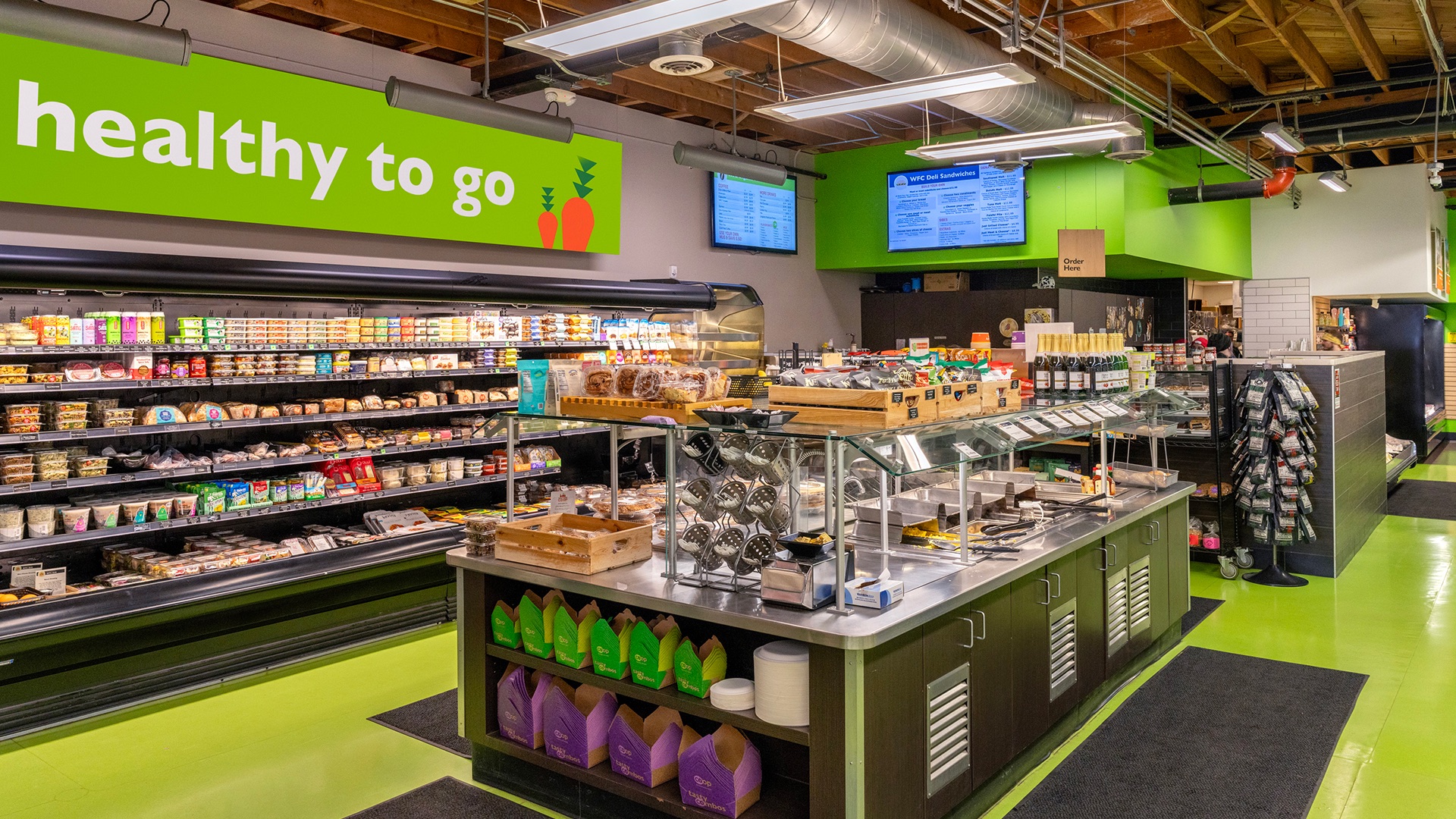 A modern grocery store section with a salad bar, shelves stocked with various food items, and a bright green sign that reads "healthy to go." The area is well-lit and features a clean design with visible food options and digital menu screens.