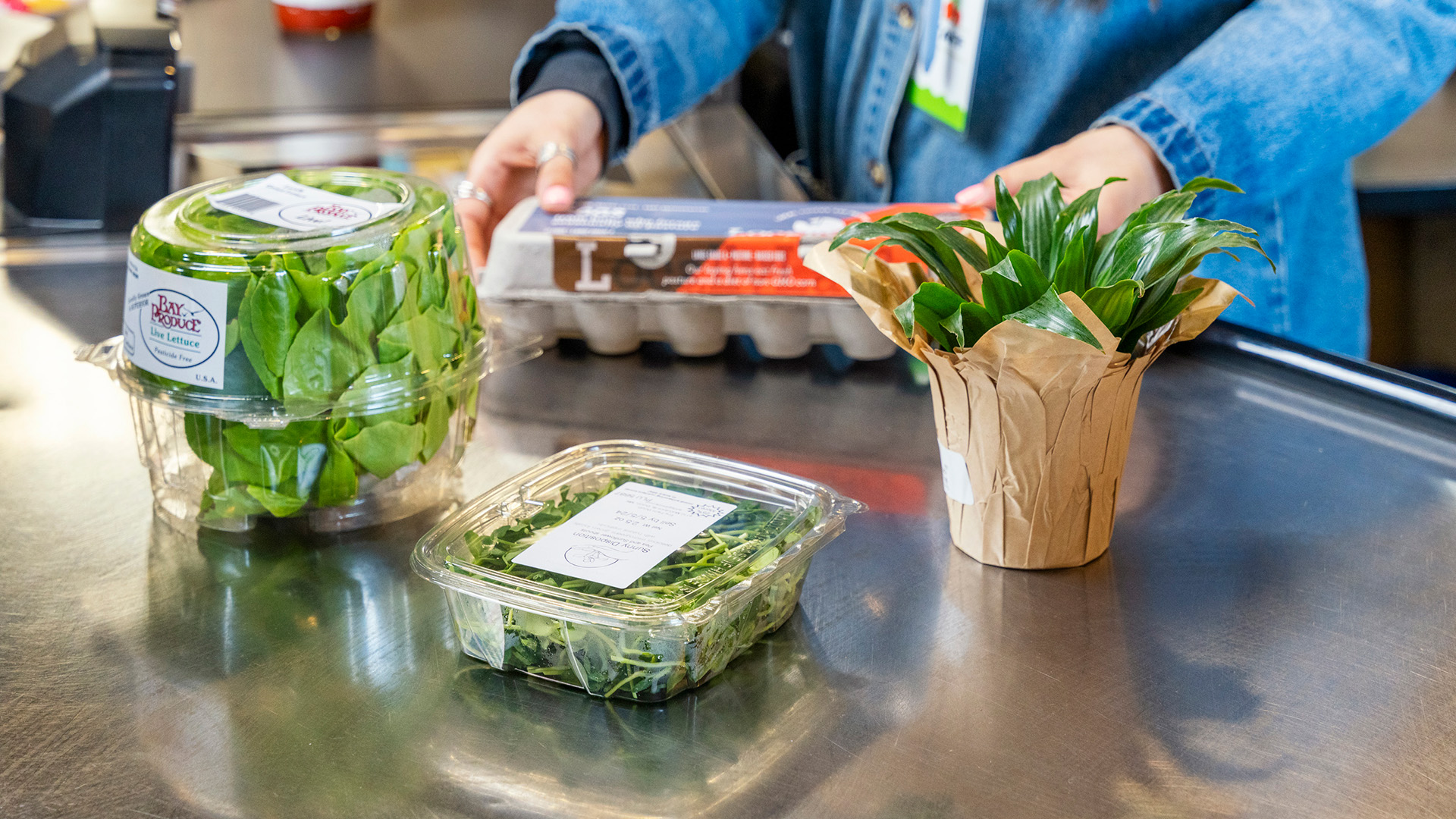 A person at a supermarket checkout places a carton of eggs, a potted plant, and two containers of leafy greens—all local produce—on the conveyor belt. The person's hands and part of a denim jacket are visible in the background.