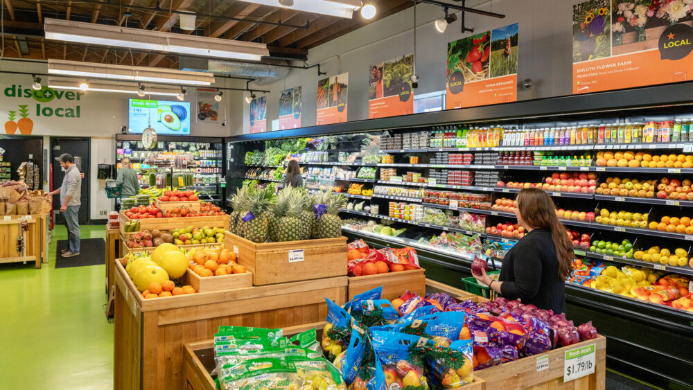 A colorful Whole Foods Coop Duluth grocery store features a wide range of fresh produce. An assortment of fruits like pineapples, melons, and apples is displayed in wooden bins, while packaged vegetables fill refrigerated shelves. Shoppers select items under bright lighting and visible "discover local" signage.