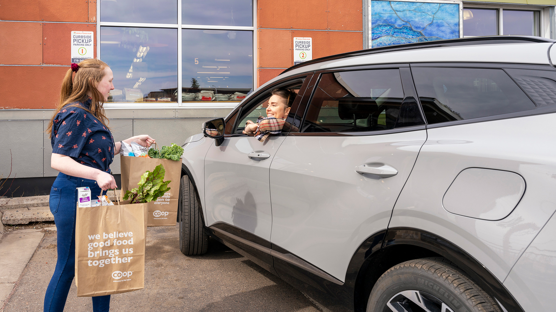 A woman at a curbside pickup location at Whole Foods Coop Duluth MN hands grocery bags to a woman in a parked car. The bags have fresh produce and a sign reading, "we believe good food brings us together." Store signs are visible in the background.