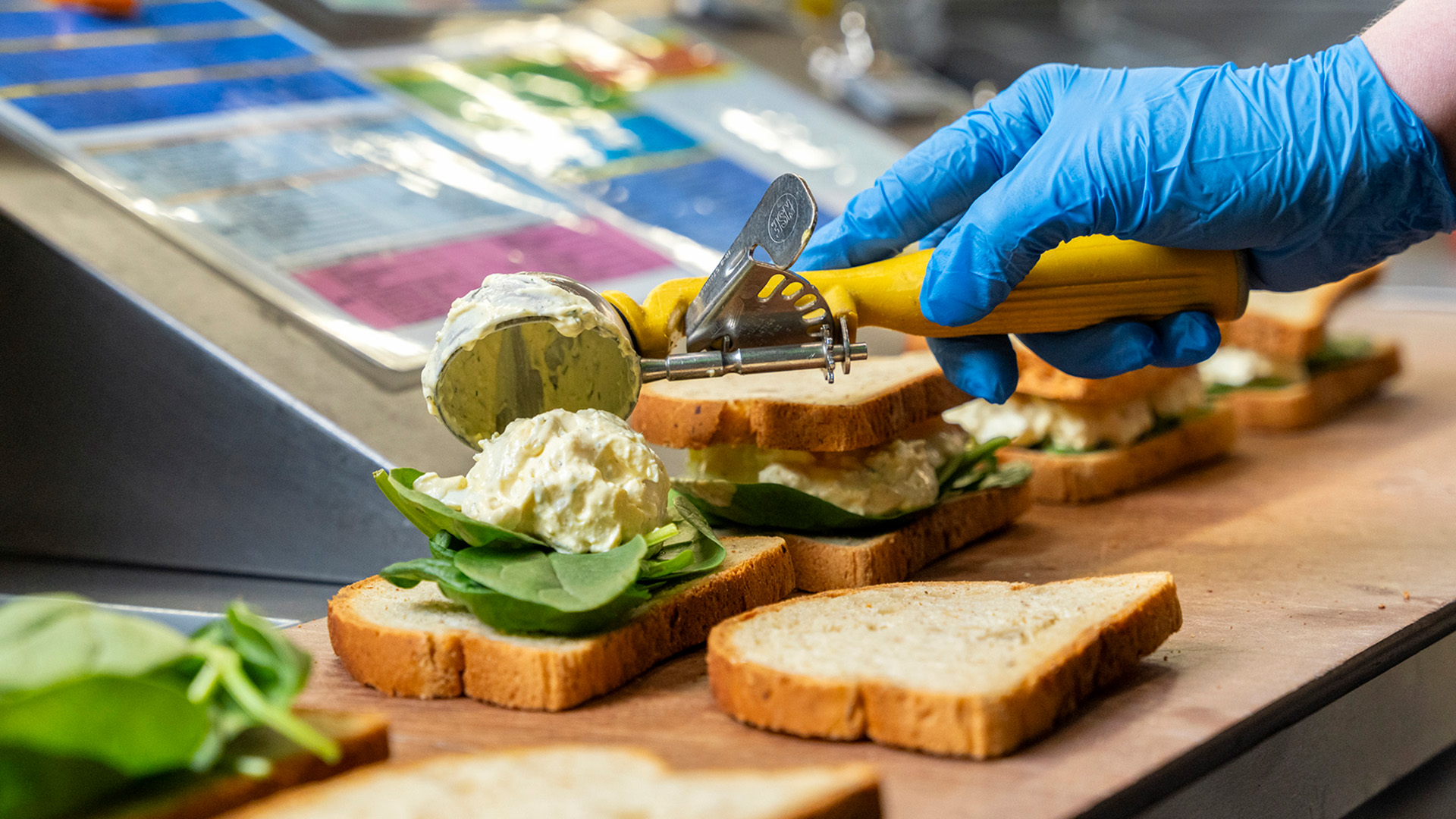 A person wearing blue gloves is using a scooper to place a dollop of sandwich filling onto slices of bread topped with spinach. The sandwiches, straight from the Whole Foods Coop Deli menu, are being prepared on a countertop, and various tools and ingredients are visible in the background.