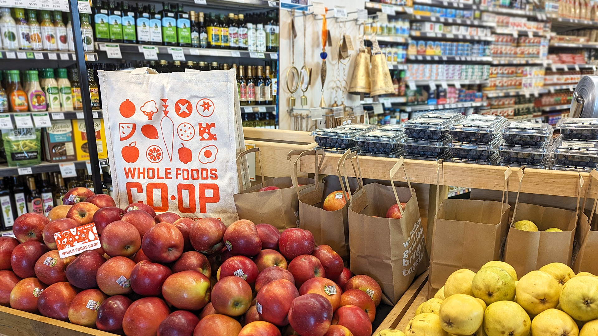 A display in a grocery store features apples and yellow apples in paper bags, and containers of blueberries. A reusable Whole Foods Coop Duluth MN shopping bag is visible among the apples, and shelves stocked with various food items are in the background.