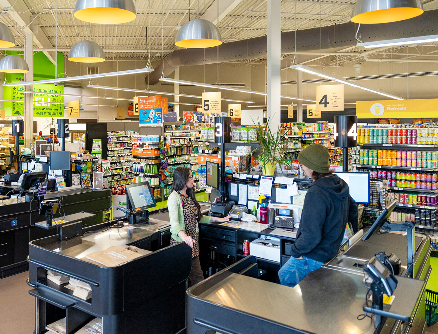 A bright, modern Whole Foods Coop checkout area in Duluth, MN features multiple conveyor belt lanes. Three cashiers are visible, each assisting different customers. Overhead lighting is contemporary, and the store's shelves are stocked with various items in the background.