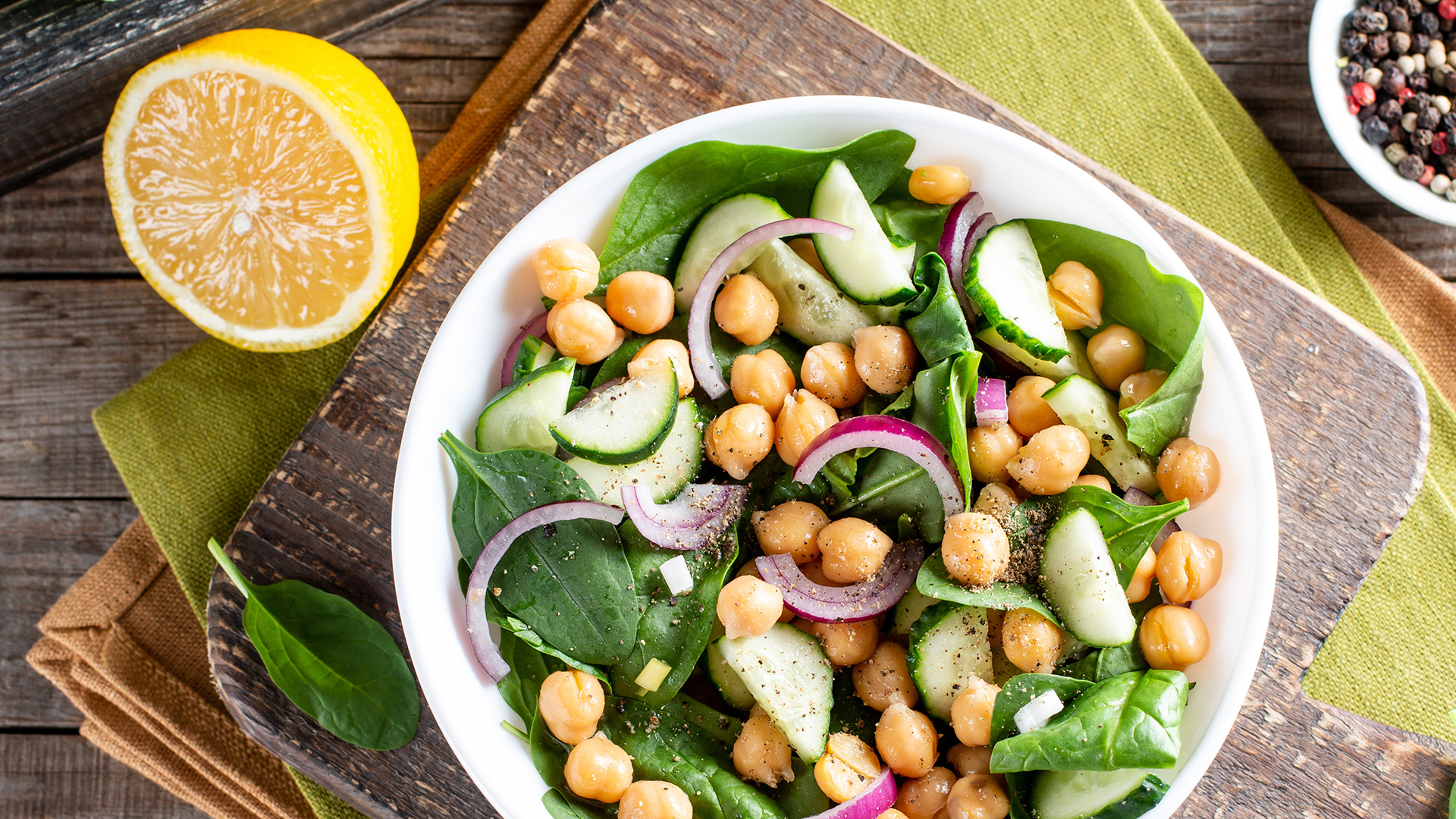 A fresh salad in a white bowl featuring chickpeas, spinach, sliced cucumbers, and red onion rings. The bowl rests on a wooden cutting board with a partial lemon and a pepper grinder visible nearby. The background includes green and brown fabric.
