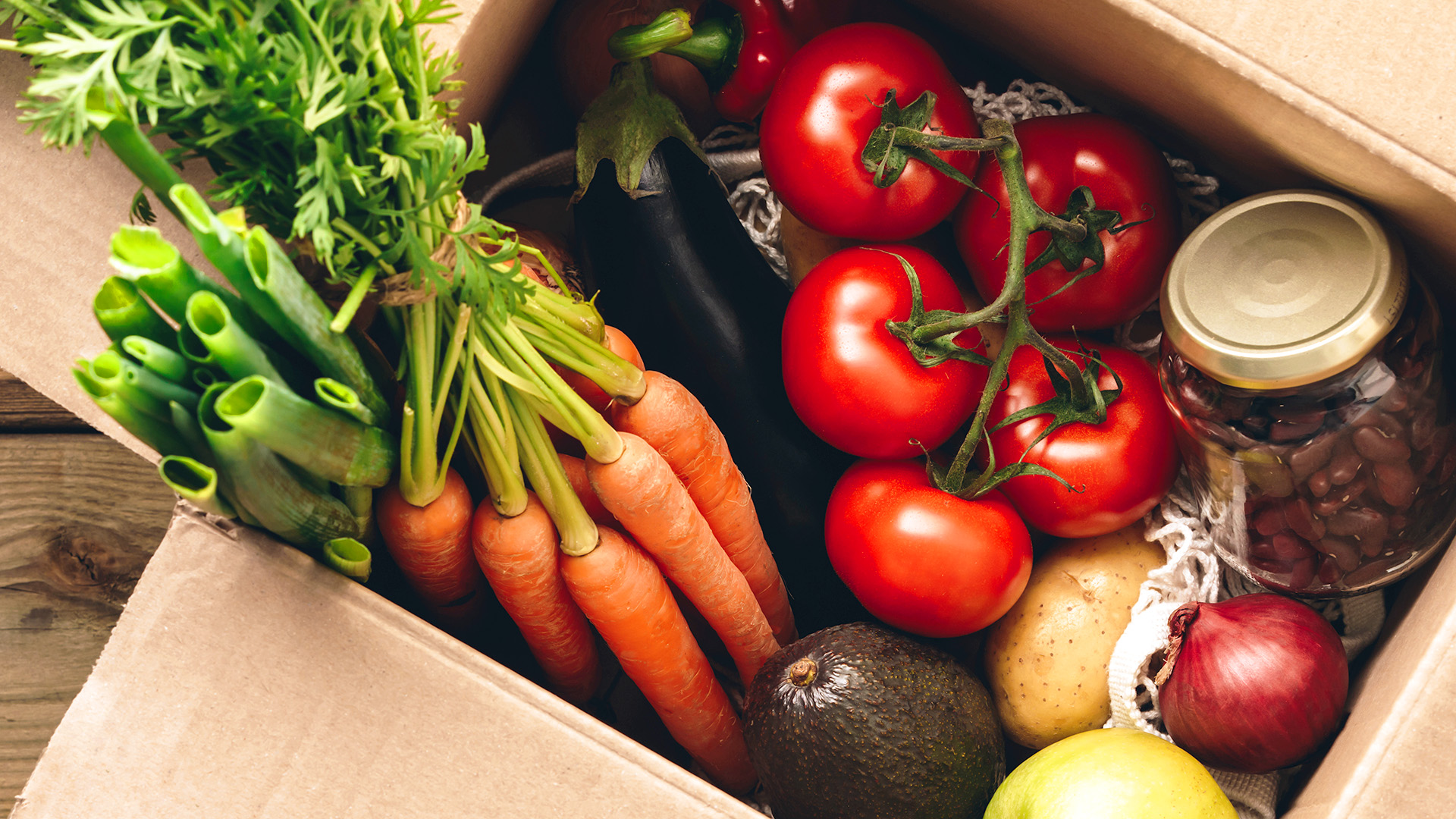 A cardboard box filled with fresh vegetables and fruit from Whole Foods Coop Duluth, including carrots with green tops, tomatoes on the vine, an eggplant, scallions, a potato, an avocado, a red onion, and a lemon. A jar of beans is also visible among the produce.