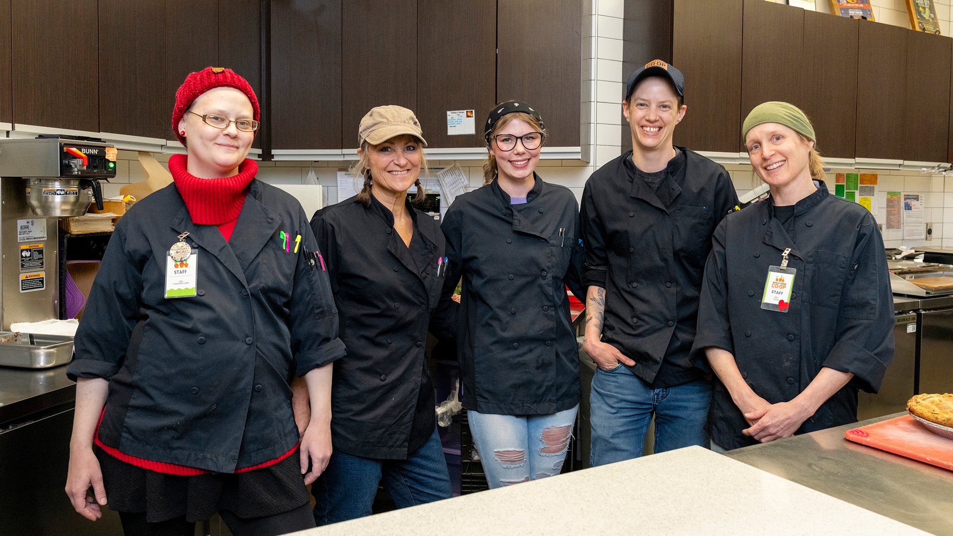 Five people wearing black chef uniforms and different colored hats stand together in a kitchen. They are smiling and standing in a relaxed, casual manner. Various kitchen equipment and utensils can be seen in the background.