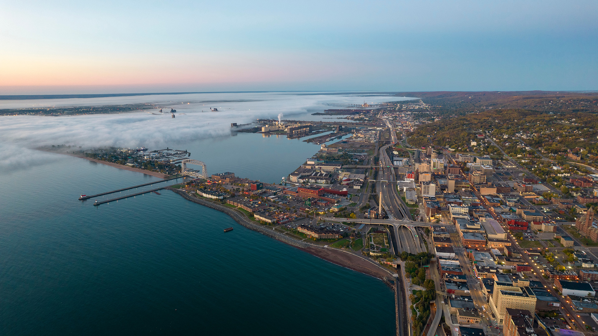 Aerial view of a coastal city with a sprawling urban area, highways, and a large body of water bordered by docks and industrial buildings. Fog covers part of the coastline in the distance. The sky is clear with soft pastel hues from the setting or rising sun near Whole Foods Co-op Duluth MN.