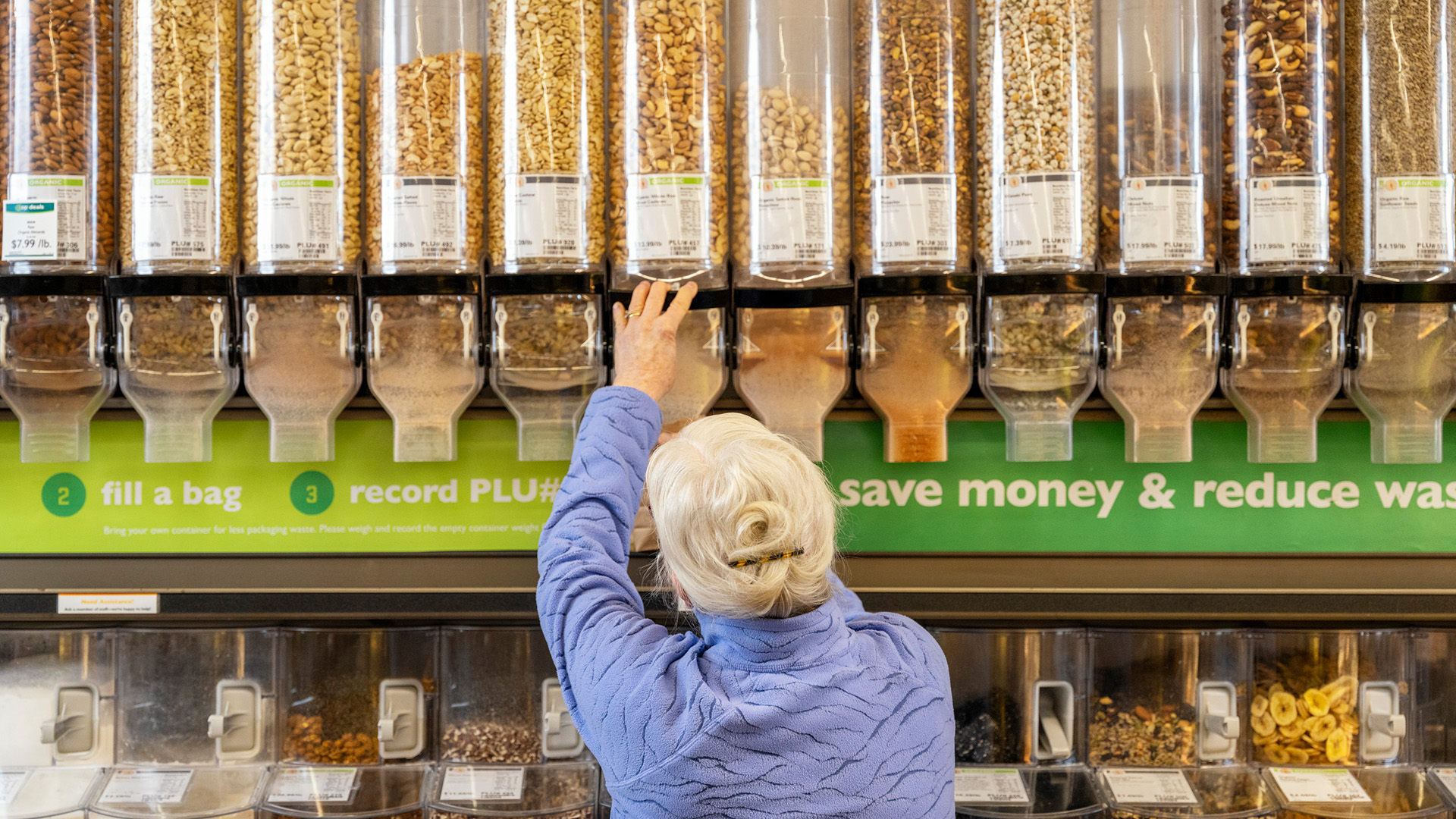 An elderly person with white hair and a blue jacket reaches for a lever on a bulk food dispenser in a store. Various grains, nuts, and cereals are visible in dispensers, with signs explaining the process of filling a bag and the benefits of reducing waste.