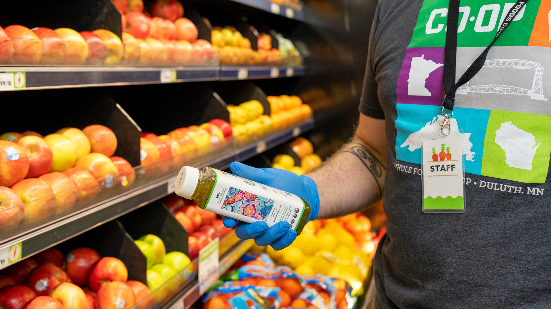 A grocery store employee wearing a "Staff" badge and blue gloves holds a bottle of green juice in front of a display of apples and bell peppers. The employee's T-shirt features the word "CO-OP" and an illustration of Minnesota, highlighting the opportunities for Whole Foods Coop jobs.