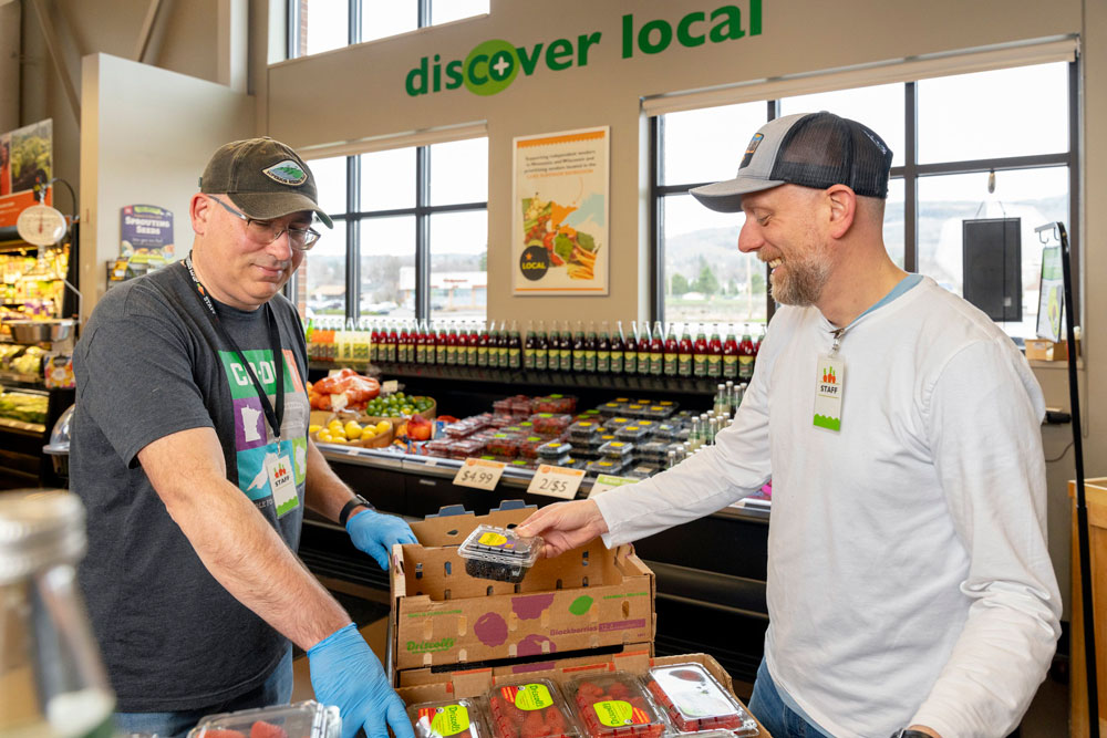 Two men are arranging fresh produce at a grocery store. One man is handing a box of blueberries to the other. They are both wearing casual clothes with hats and name tags. The background shows shelves with various fruits and a sign that says 