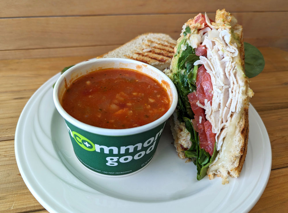 A cup of tomato soup and a turkey sandwich with spinach, tomato, and avocado on toasted bread are served on a white plate. The soup cup has 