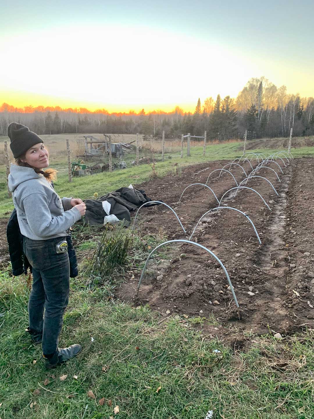 A person wearing a beanie, hoodie, and jeans stands next to a freshly prepared garden bed with metal hoops under a sunset sky. The garden bed is ready for planting, and there's a rustic fence and trees in the background. The person is smiling and holding some small objects.