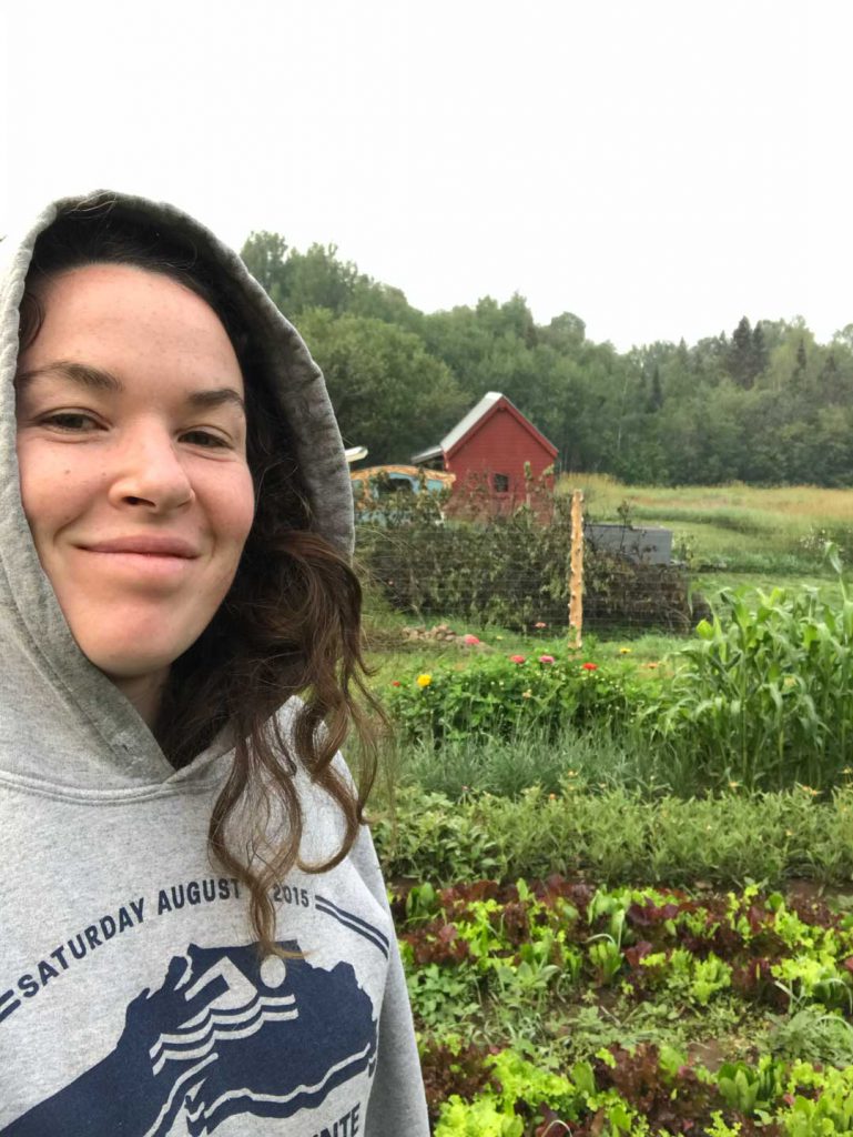A person wearing a hooded sweatshirt stands in a garden with various plants. They are smiling with trees and a red cabin visible in the background. The sweatshirt has a design with text partially visible, featuring the date 