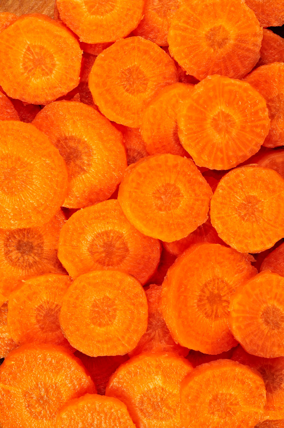 A close-up image of numerous carrot slices, showing their vibrant orange color. The slices are evenly cut and some have visible core rings in the center, creating a textured and layered appearance.