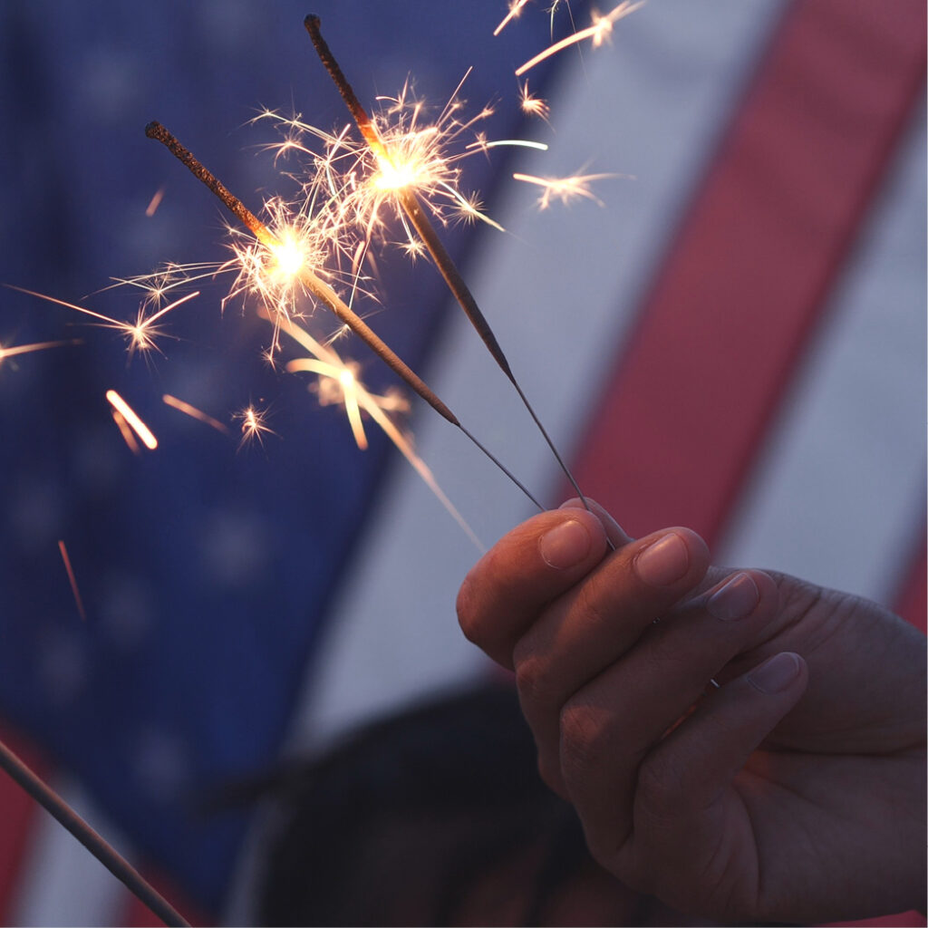 Close-up of a hand holding three lit sparklers against a blurred background featuring a partially visible American flag. The sparkling light from the sparklers contrasts with the dark and out-of-focus backdrop, creating a sense of 4th of July celebration.