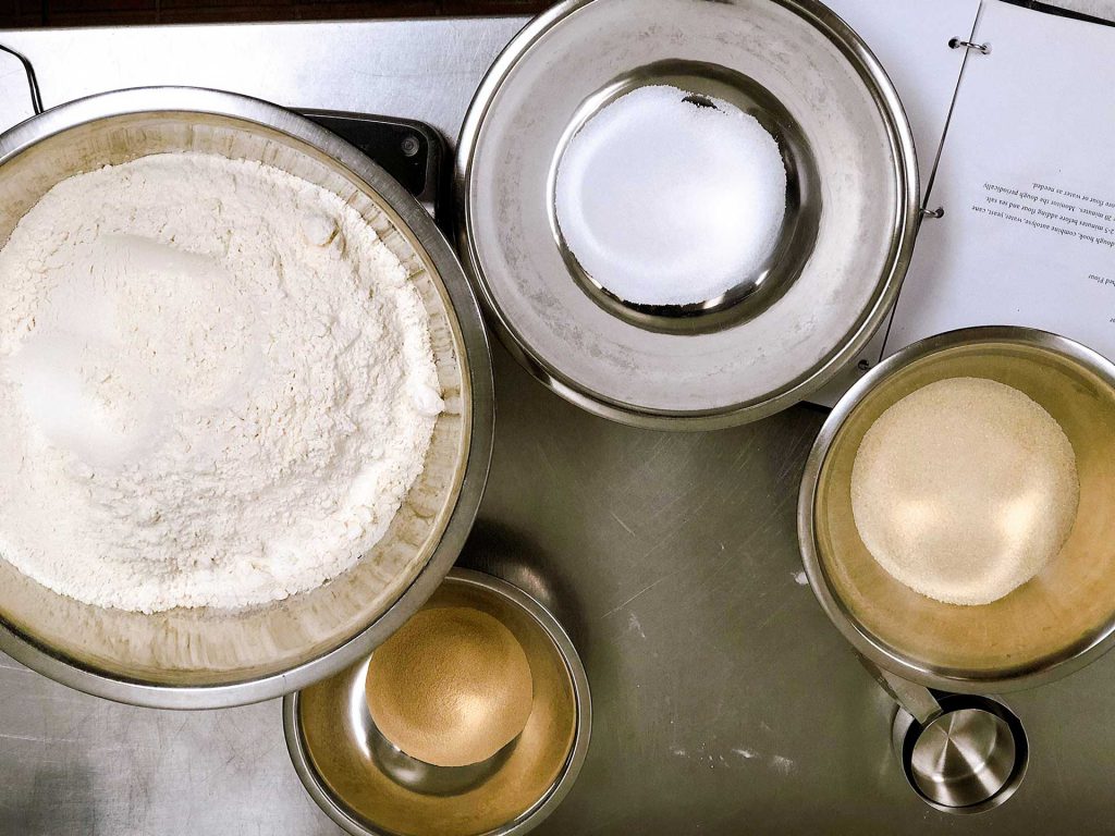 Four metal bowls containing different dry ingredients are arranged on a stainless steel surface. The largest bowl holds flour, while the smaller bowls contain sugar and possibly other powders. An open book or recipe is partially visible to the right.