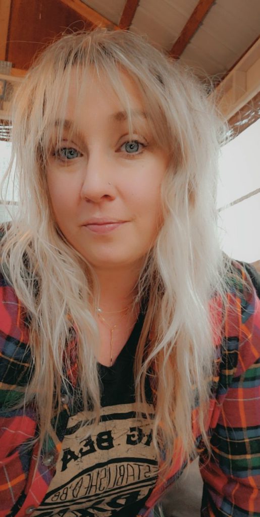 A woman with wavy, long blonde hair and blue eyes is looking at the camera. She is wearing a red and green plaid shirt over a black top with a graphic print. The background includes a wooden ceiling and part of a window, suggesting an indoor setting.
