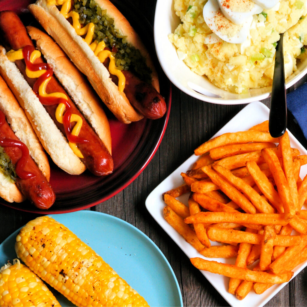 A delicious Labor Day spread featuring three hot dogs with mustard and relish on a plate, a bowl of creamy potato salad with an egg on top, a plate of crispy seasoned fries, and two ears of grilled corn on a blue plate, all set on a dark wooden table.