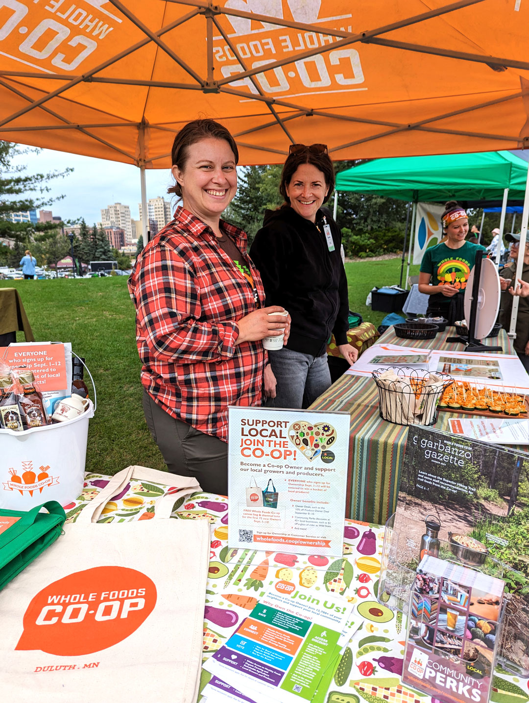 Two people stand behind a table at an outdoor event booth for Whole Foods Coop Duluth MN. The table displays brochures and promotional materials. They smile at the camera, with one person in a red plaid shirt and another in a black jacket. A tent and trees are in the background.