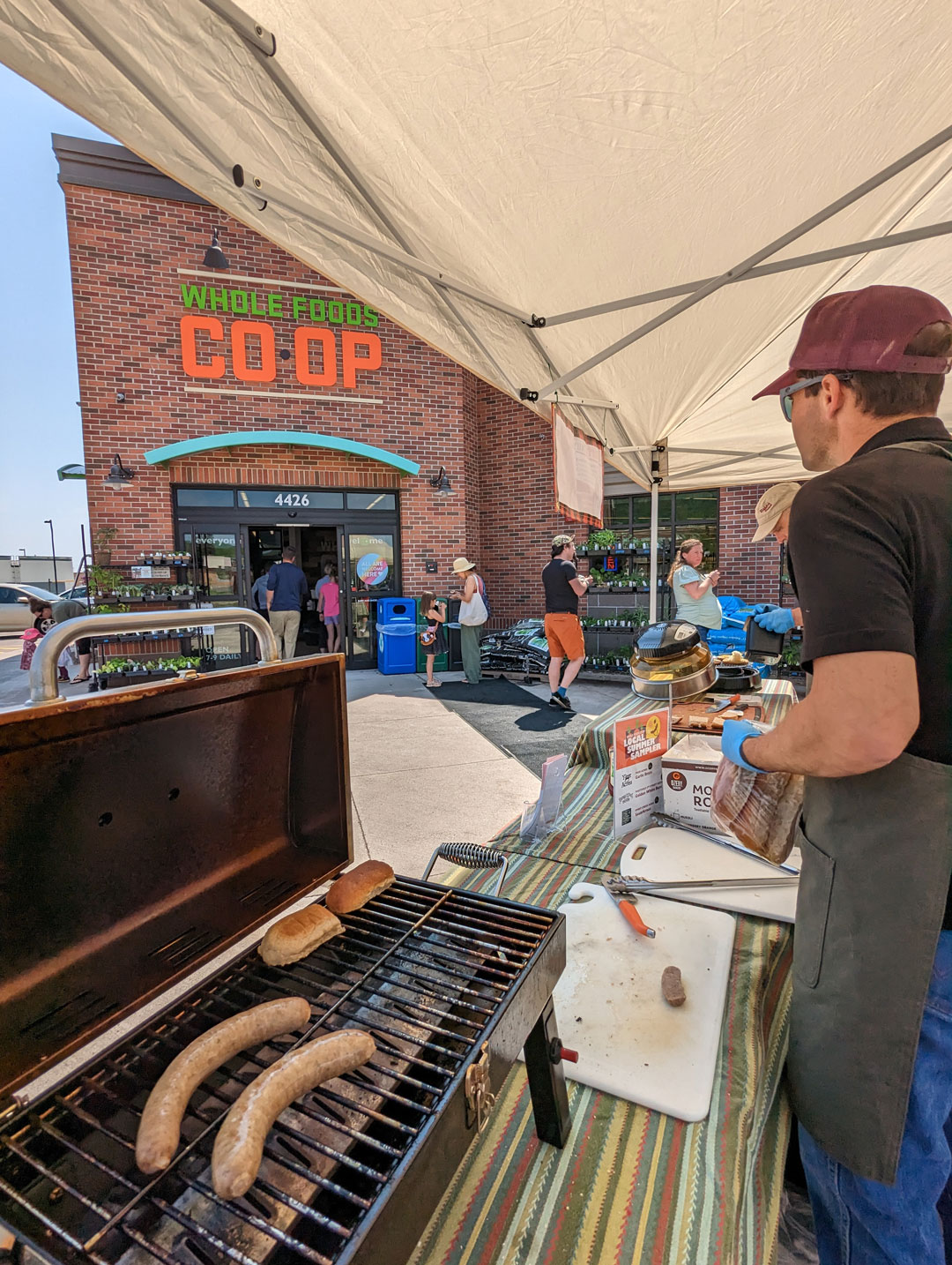 A vendor cooks sausages on a grill under a tent outside the Whole Foods Co-op Duluth MN store. Several people are gathered around, some shopping and others talking. The storefront is brick, and there is a sign reading "WHOLE FOODS CO OP" above the entrance.