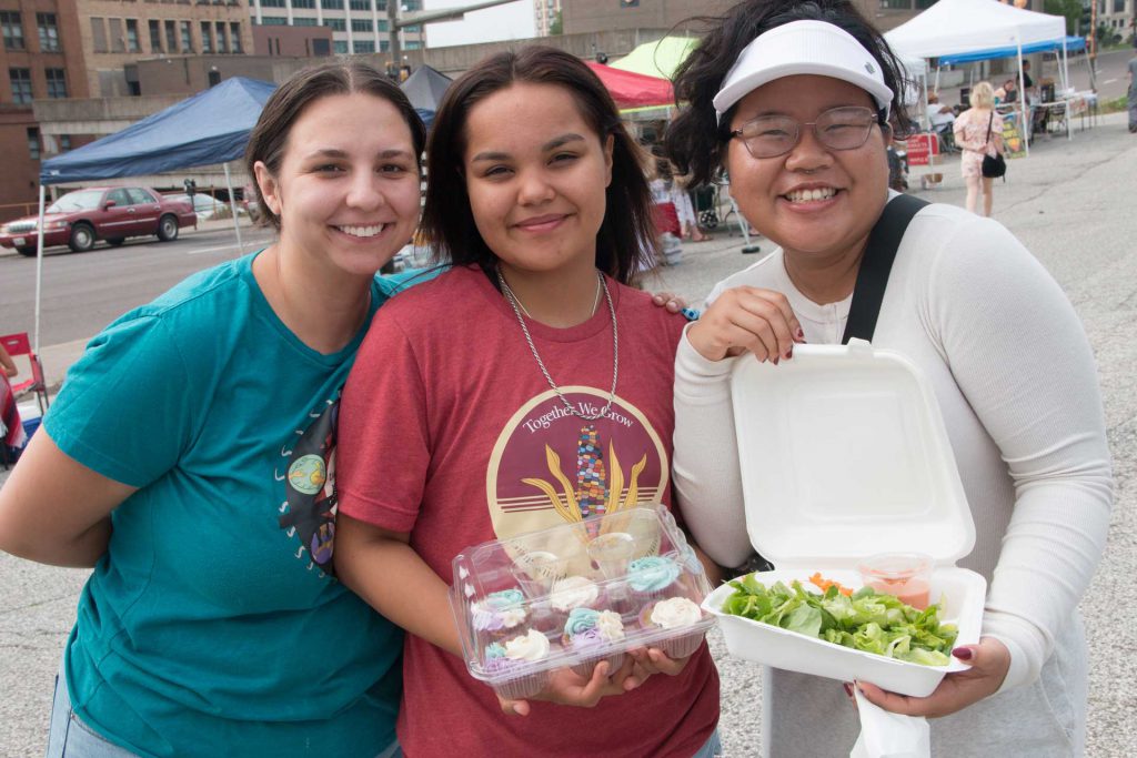 Three people are smiling at an outdoor market. The person on the left is wearing a blue shirt, the one in the middle is in a red T-shirt holding a box of cupcakes, and the person on the right is in a white shirt and hat holding an open container with a salad.
