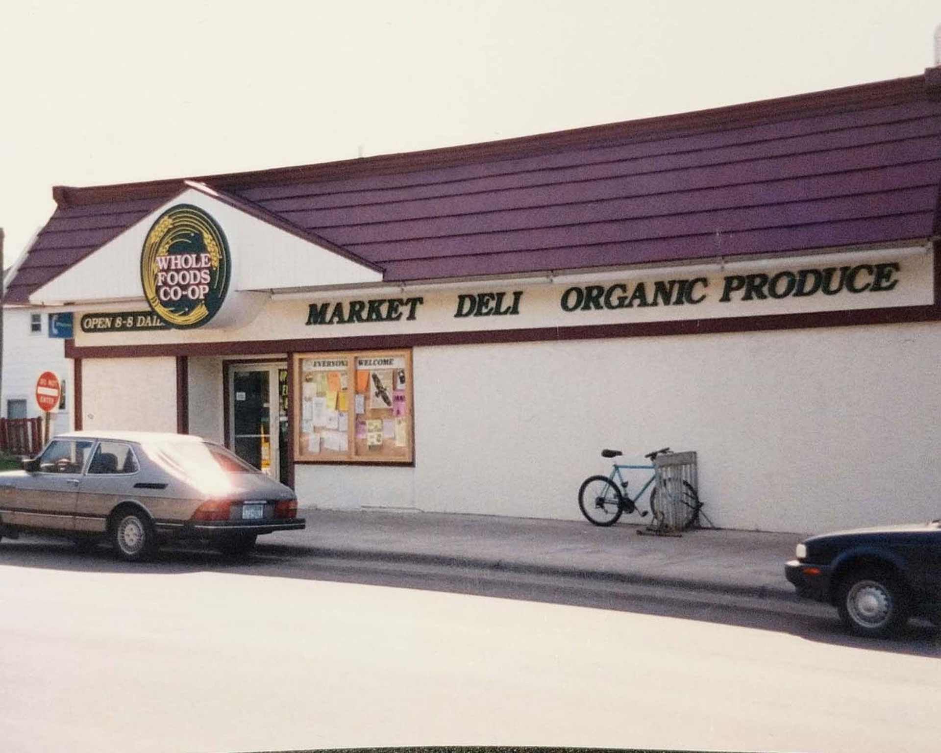 A vintage photo of a Whole Foods Co-op store in Duluth, MN with a maroon-roofed building. Signs read 