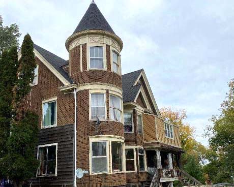 A multi-story, old-style brick house with a distinctive round turret featuring multiple windows stands proudly near the Whole Foods Coop in Duluth, MN. The house has a dark shingle roof and is surrounded by trees and greenery. A set of stairs leads up to the front porch under an overcast sky.