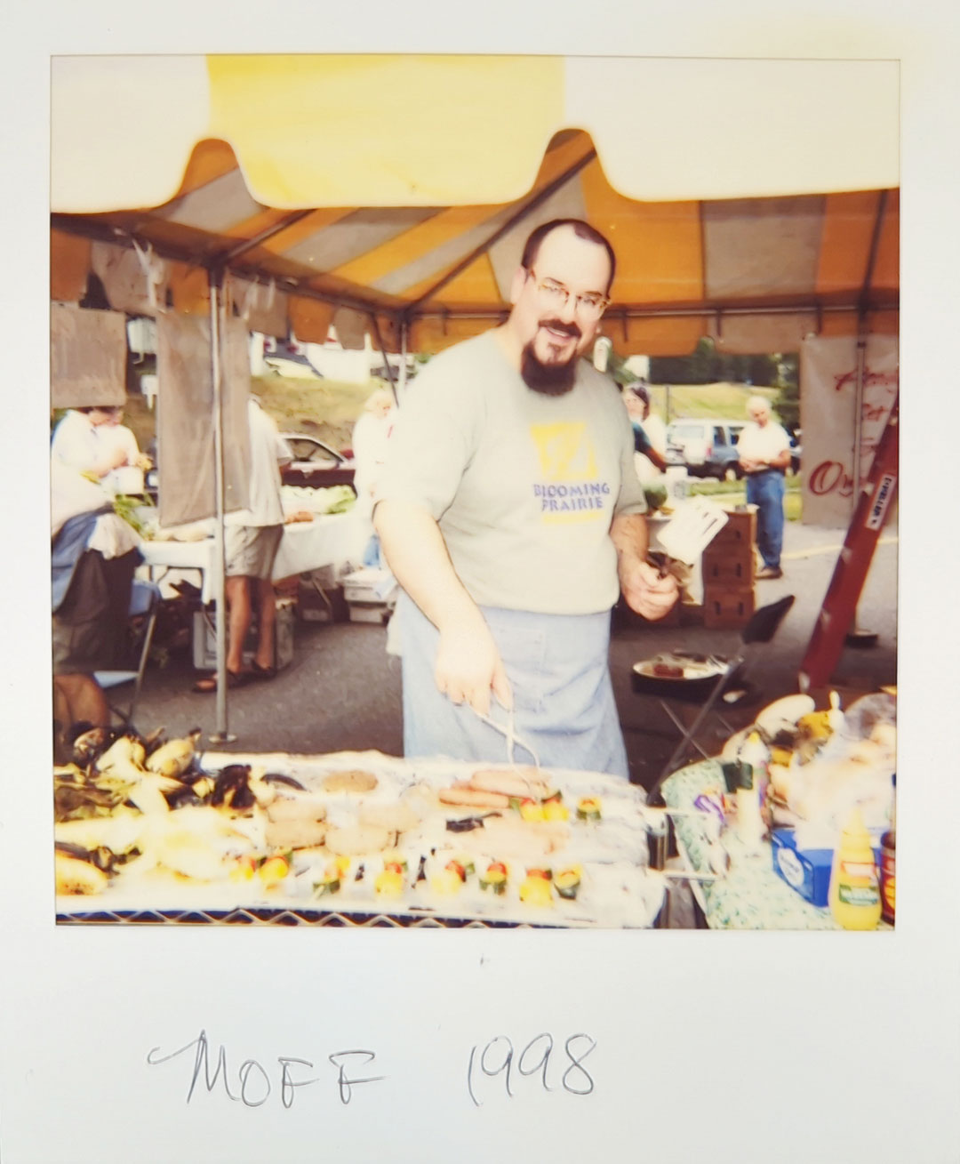 A person smiling while grilling food at an outdoor market stall. The person, wearing a t-shirt and apron, is holding a grilling fork, and various food items can be seen cooking on the grill. A yellow and white canopy covers the stall at Whole Foods Coop Duluth MN. Handwritten text below the image reads "MOFF 1998.