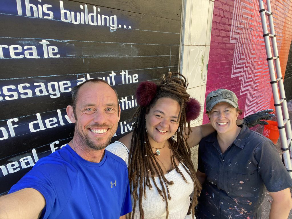 Three people standing in front of a colorful mural that features text and geometric patterns. On the left, a person wearing a blue shirt smiles at the camera. The person in the middle has dreadlocks and wears a white top. The person on the right is in a dark shirt and cap.