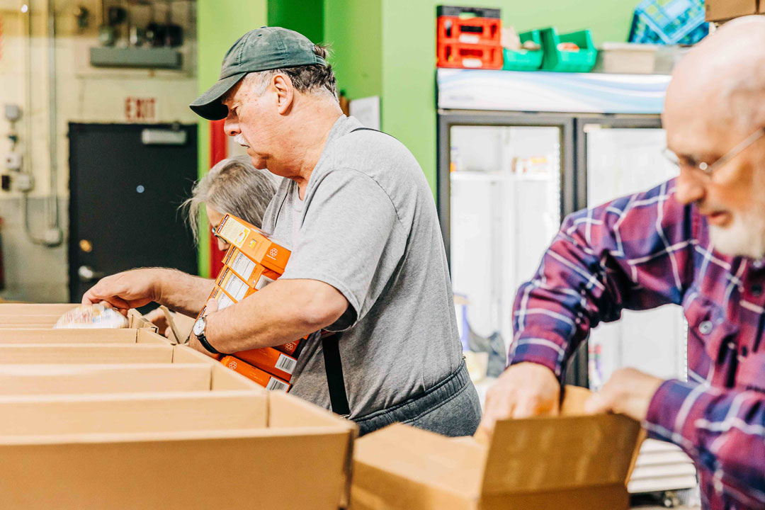 Three older adults assemble food boxes in a brightly lit setting at Whole Foods Coop Duluth. One man in a gray shirt and black cap holds a box of cereal, while a woman in a similar shirt and another man in a plaid shirt work alongside him. Shelves and a refrigerator are visible in the background.
