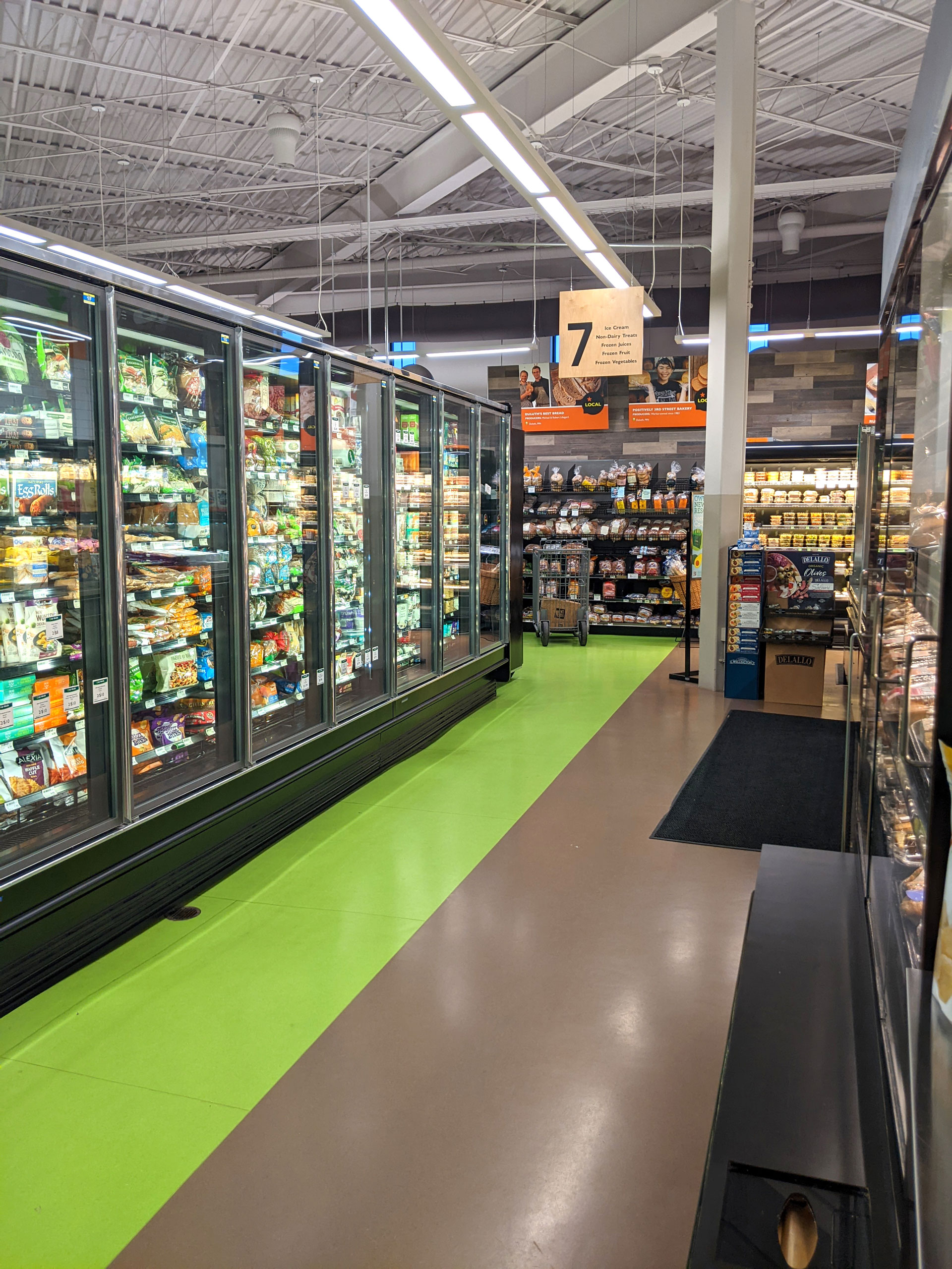 A supermarket aisle with refrigerator units displaying various frozen foods on the left and bakery items or snacks on the right. The brightly lit space has green and brown flooring, and a sign overhead marking aisle 7.