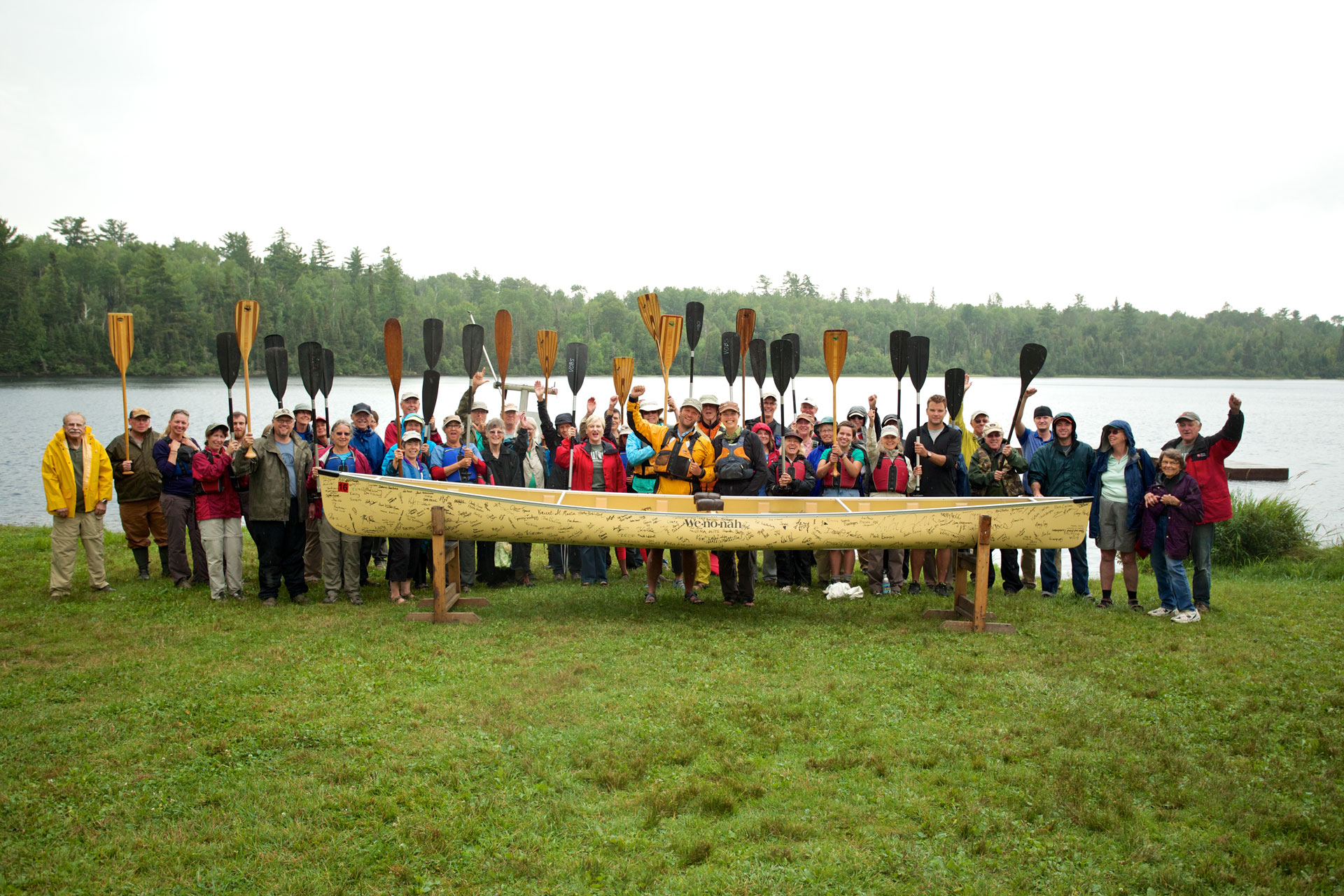 A large group of people, dressed in outdoor gear, stand in front of a lake holding paddles raised in the air, with a canoe placed on stands in front of them. The background features a forested shoreline under an overcast sky.