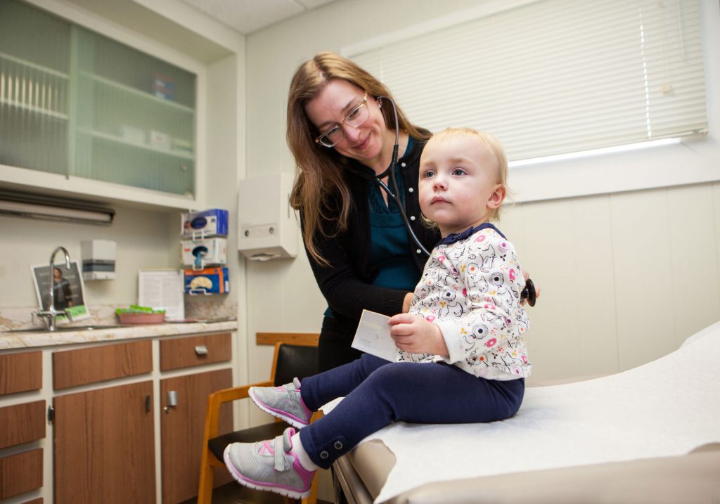 A healthcare professional, wearing glasses and a black cardigan, smiles while attending to a young child sitting on an examination table in a medical office. The child is wearing a floral-patterned shirt, blue pants, and pink shoes. The room has cabinets and medical equipment.