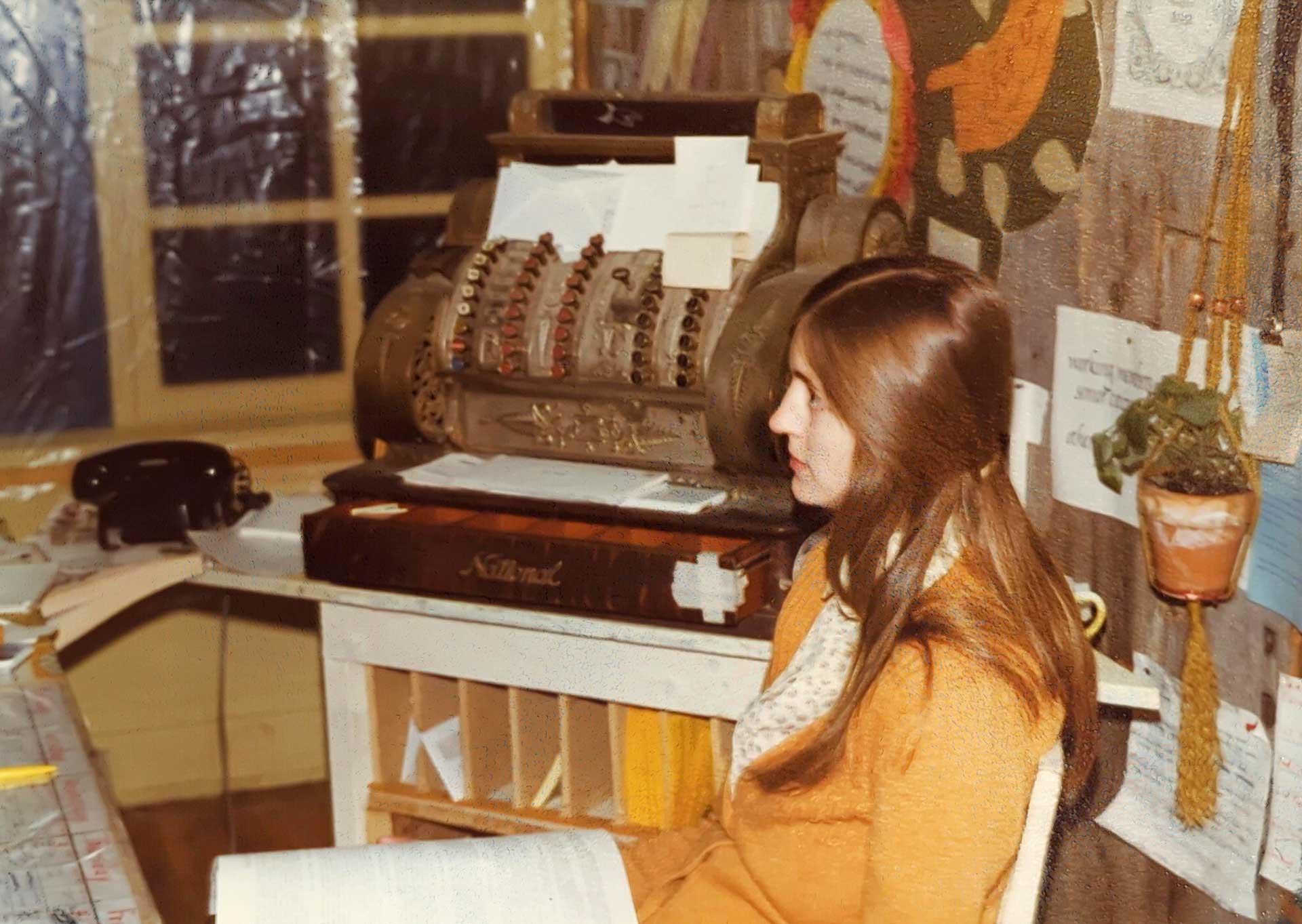 A woman with long brown hair, wearing a mustard-colored outfit and scarf, sits beside an antique cash register. The background includes a telephone, various papers, and a potted plant hanging on the wall. The setting gives off vibes of a vintage office or shop reminiscent of the Whole Foods Coop Duluth MN.
