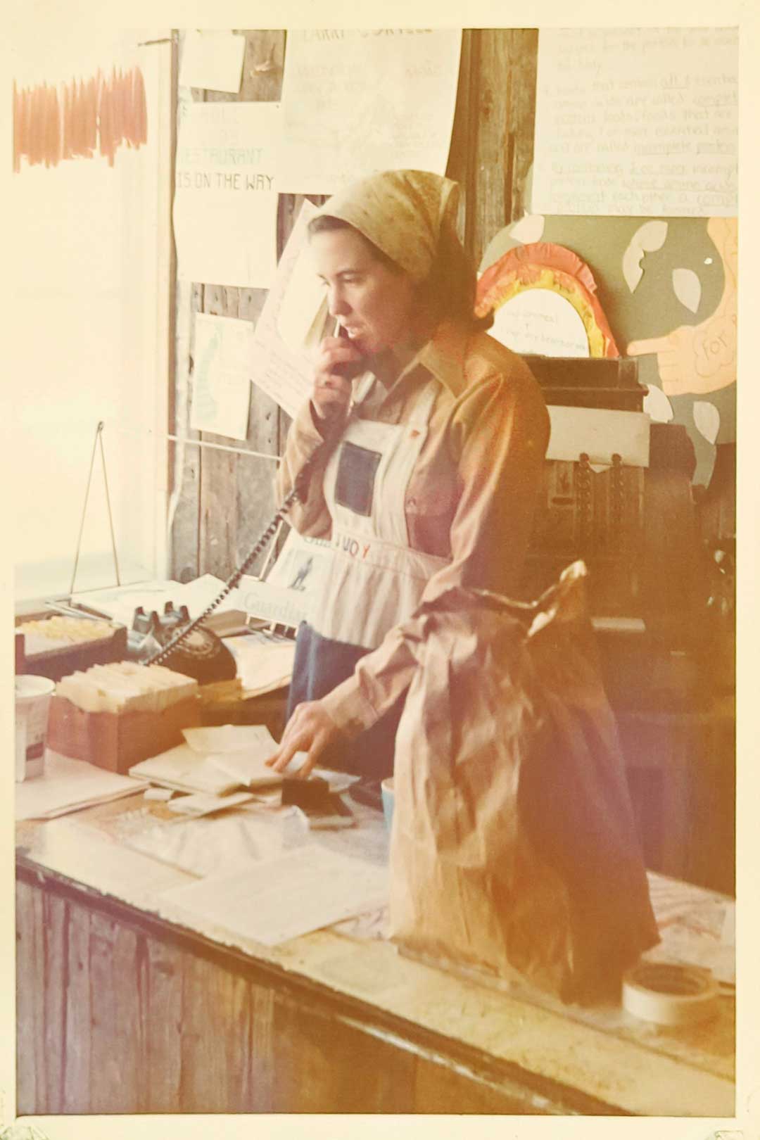 A person wearing a headscarf and apron stands at a wooden counter, speaking on a rotary phone. The counter has a brown paper bag, papers, and a cash register. In the background of this Whole Foods Coop in Duluth MN, posters adorn the walls while natural light streams through the window.
