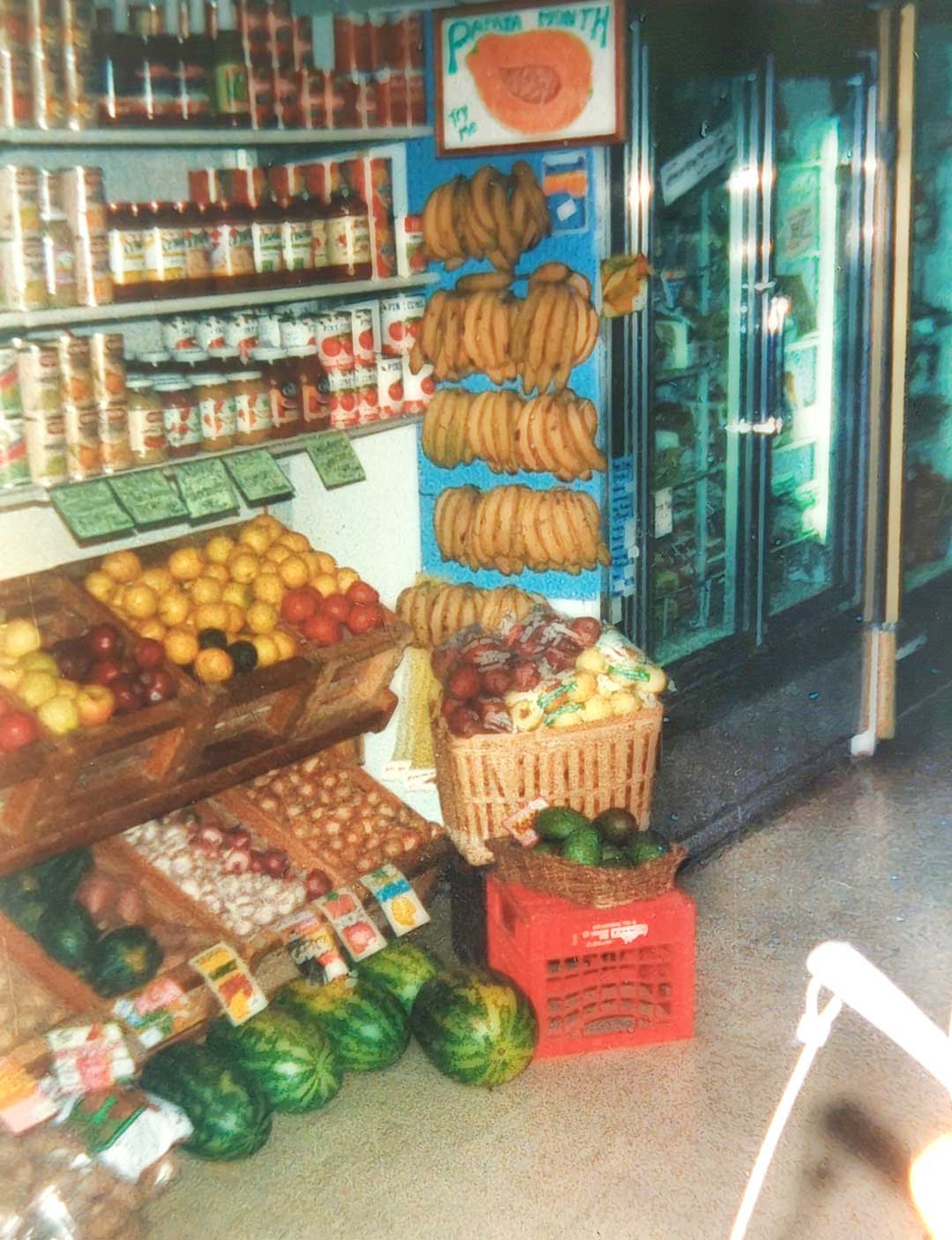 A small grocery store display at Whole Foods Coop Duluth MN features a variety of fresh fruits and vegetables. Bananas hang from a rack, while apples, lemons, watermelons, and other produce are neatly arranged in baskets and crates. Shelves are stocked with canned goods and jars.