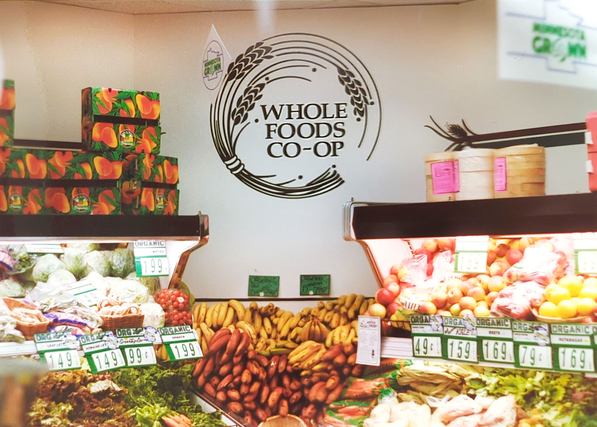 A colorful supermarket produce section featuring fresh fruits and vegetables displayed on shelves. A "Whole Foods Co-Op Duluth MN" sign is prominently visible on the wall. Price tags indicate a variety of produce including bananas, apples, and leafy greens.