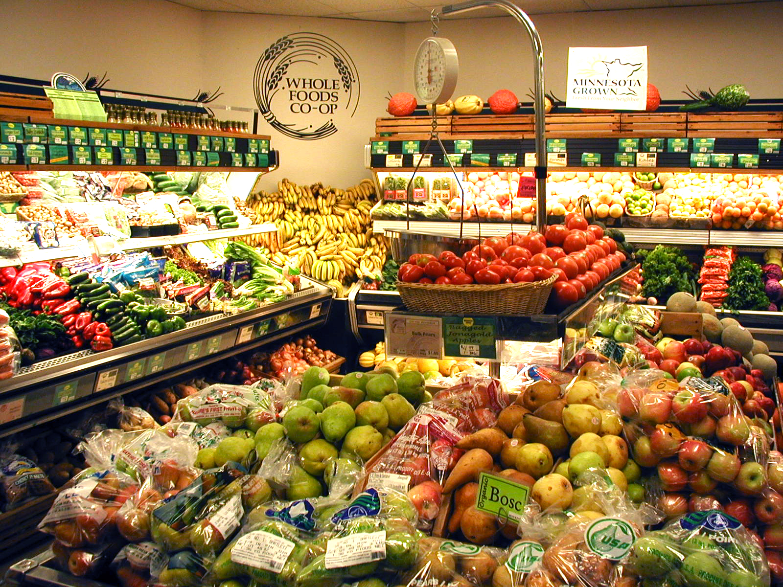 A well-lit grocery store produce section. Shelves are filled with a variety of fresh vegetables and fruits, including bananas, tomatoes, apples, pears, and bags of produce. Signs reading "WHoLE FOODS CO•OP Duluth MN" and "MINNESOTA GROWN" are displayed above the shelves.