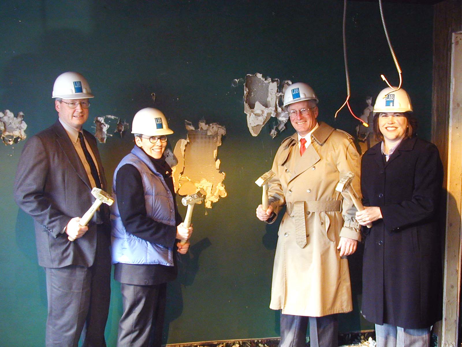 Four people wearing construction helmets pose with hammers in front of a wall with exposed studs and broken drywall. Dressed in business attire, one sports a trench coat and another a blue vest, giving the scene of this Whole Foods Coop Duluth MN event a unique blend of professionalism and construction spirit.