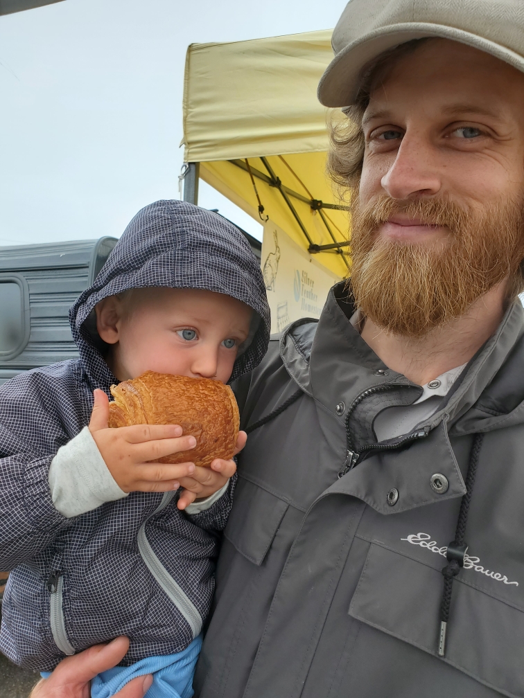 A man with a beard and a baseball cap holds a young child wearing a gray hooded jacket. The child is eating a large piece of bread or pastry. They are outdoors near a covered market stall with a yellow canopy.