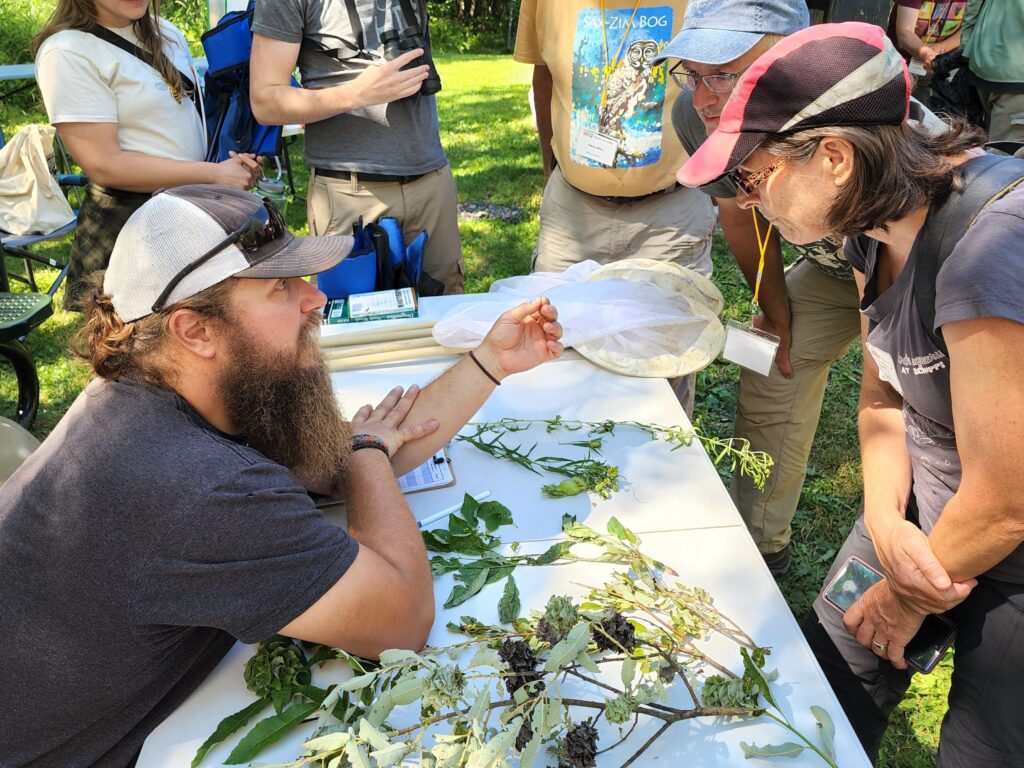 A bearded man sits at a table covered with various plants, holding up a sample in a plastic bag. He is explaining something to a woman leaning in closely, while others stand around observing. The scene takes place outdoors in a park-like setting.