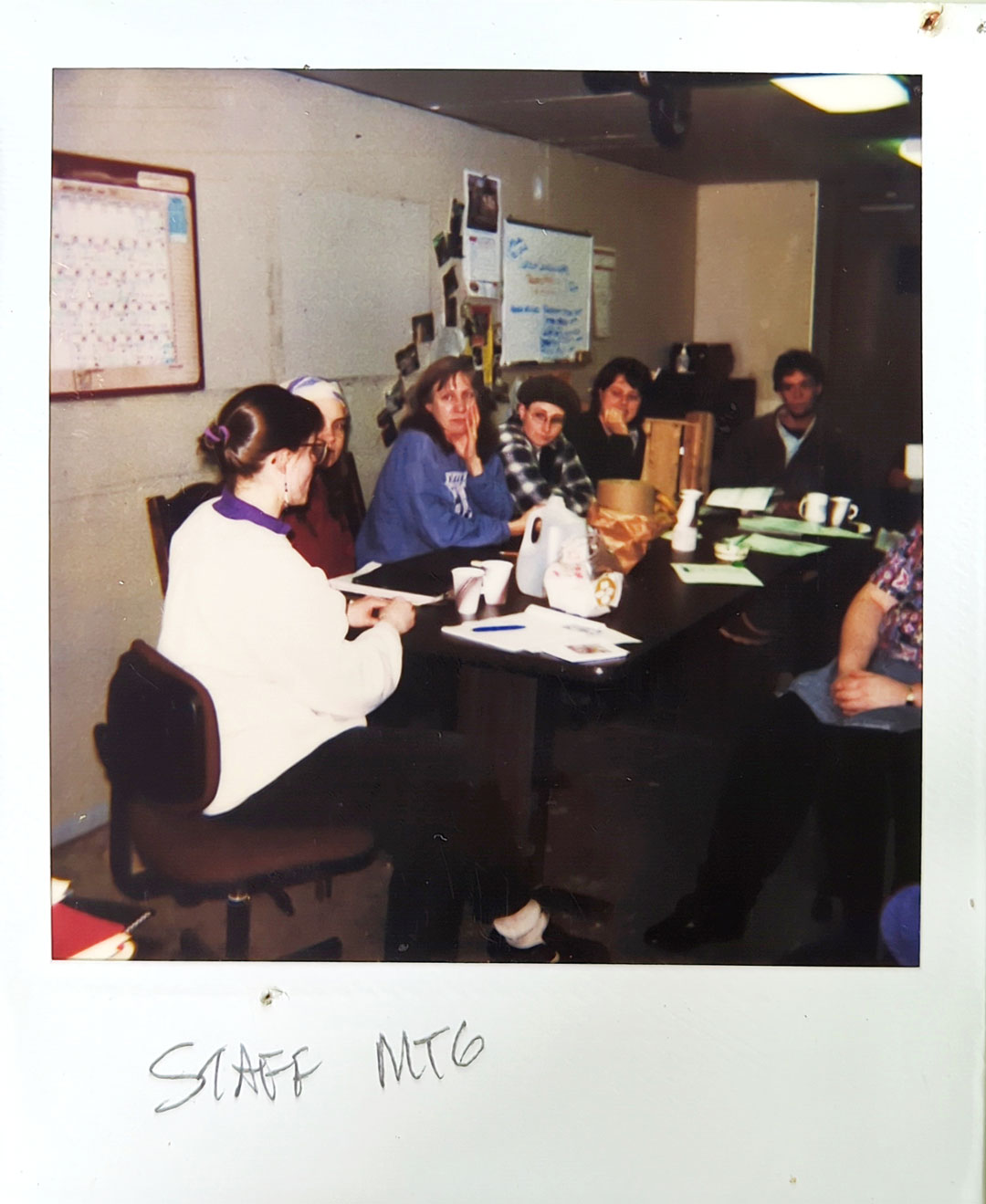 A group of six people is seated around a table in a small room for a meeting. Papers, a coffee maker, and cups are on the table. A handwritten note below the image reads "STAFF MTG." At Whole Foods Coop Duluth MN, a calendar and papers are pinned on a bulletin board in the background.